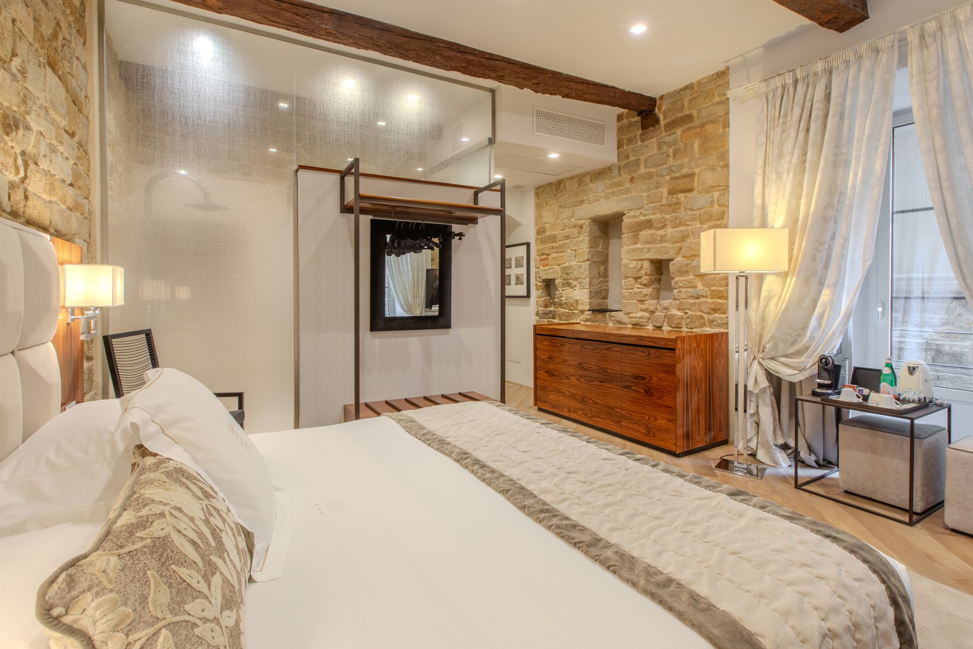 Modern hotel bedroom with exposed stone walls, a large bed with neutral bedding, a wooden sideboard, a floor lamp, and a minimalist open wardrobe.