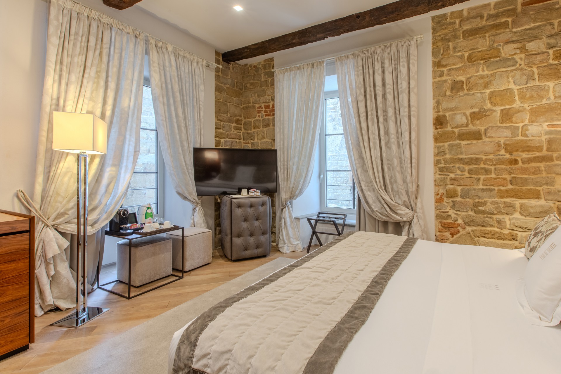 A spacious hotel bedroom featuring exposed stone walls, wooden ceiling beams, two large curtained windows, a bed with cream linens, and a flat-screen TV.