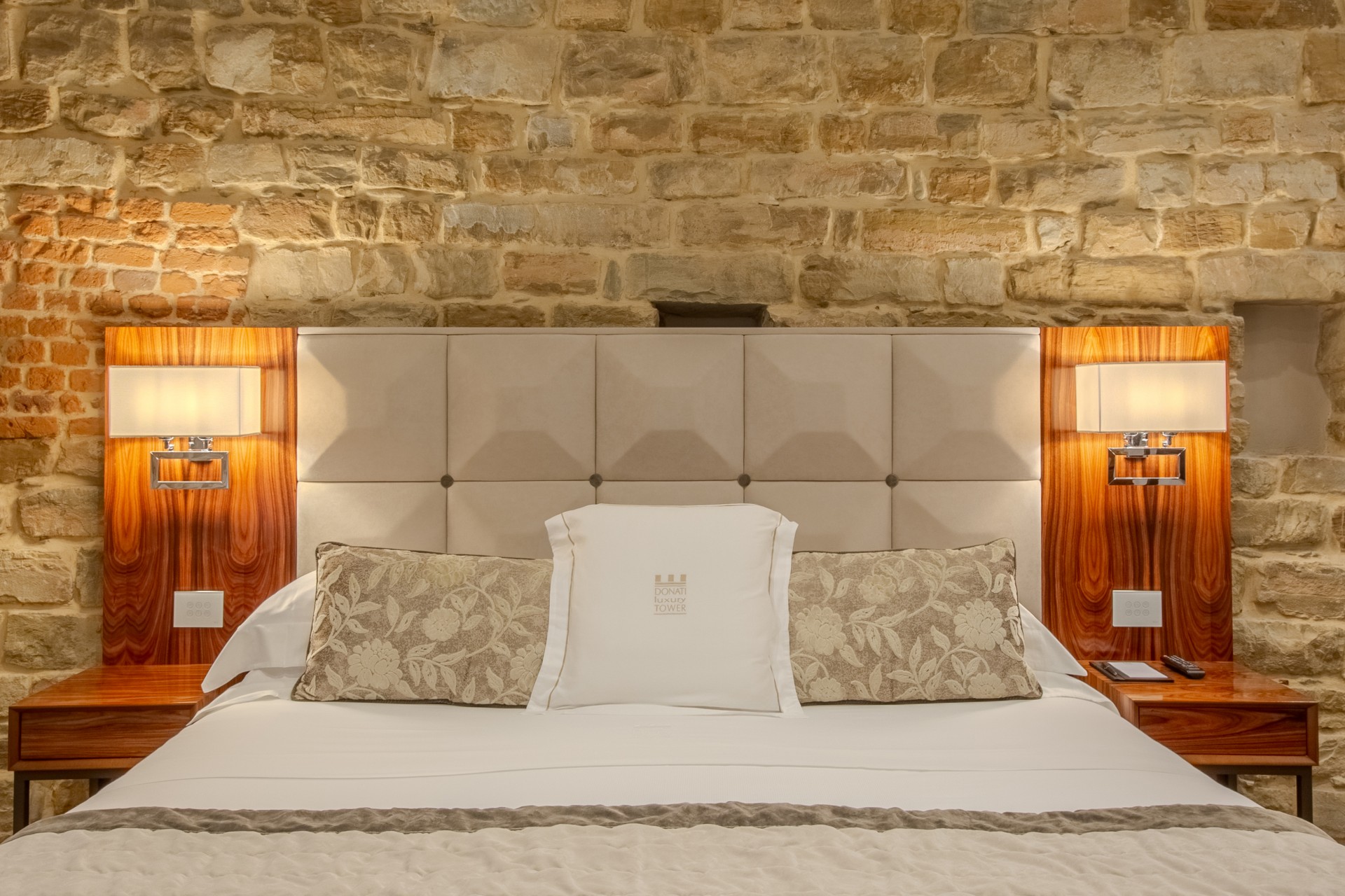 Close-up of a hotel bed set against an exposed stone and brick wall, featuring a beige tufted headboard, wooden side panels with mounted lamps, and matching nightstands.