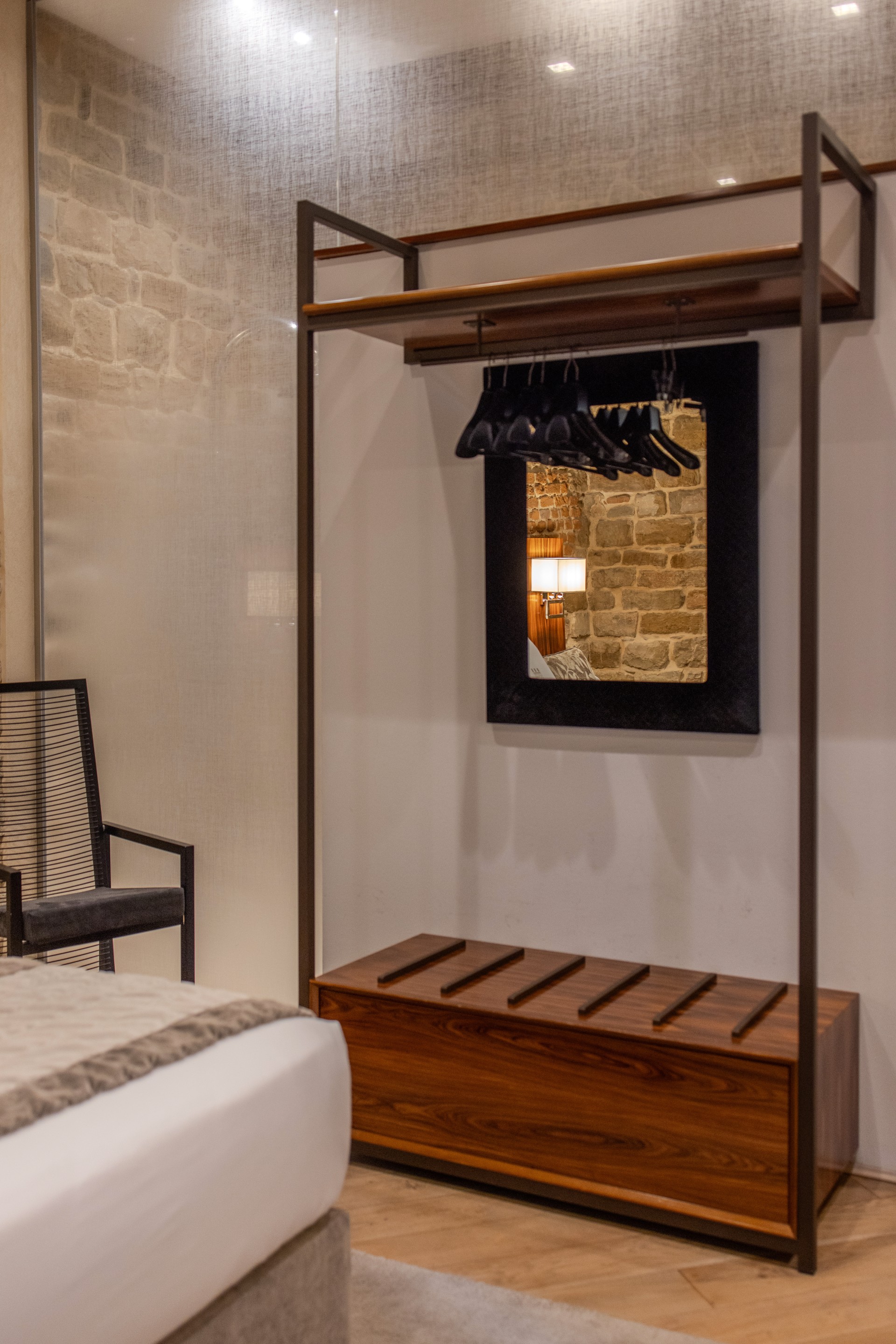 Modern open closet unit in a luxury hotel room, featuring dark metal framing, a wooden luggage rack base, coat hangers, and a black-framed mirror reflecting an exposed stone wall.