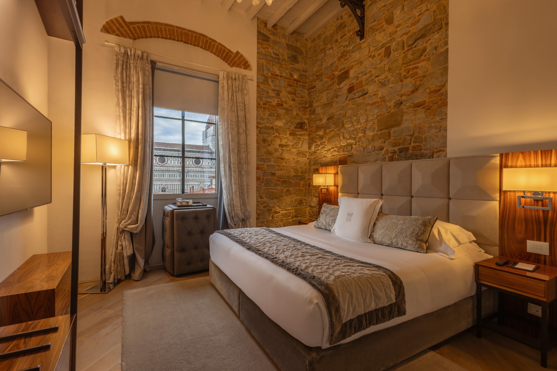 Spacious hotel bedroom with a plush king-size bed, warm bedside lighting, an exposed stone wall, wooden credenza, and a window framing a view of historic Florence architecture.