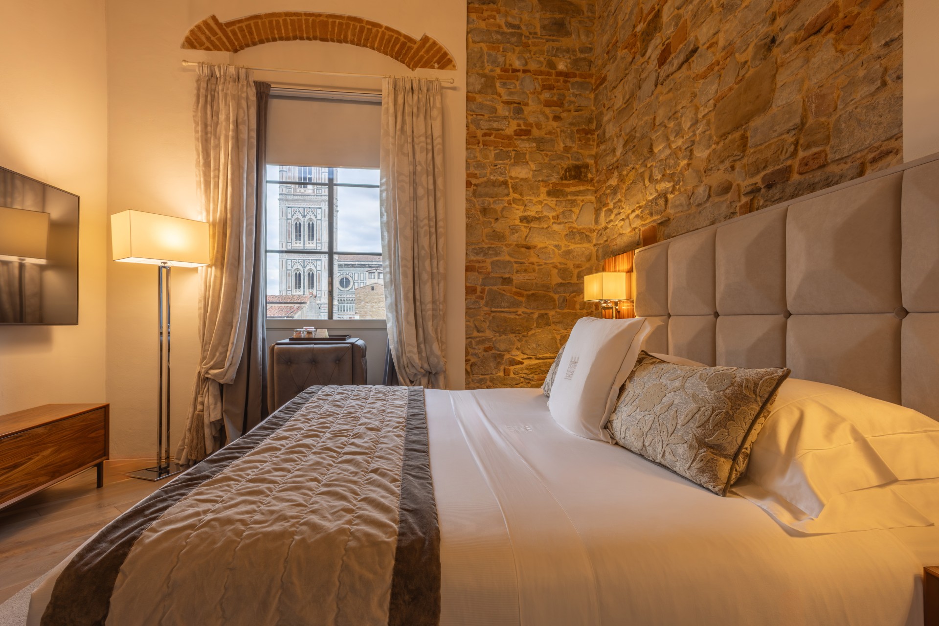 Cozy hotel bedroom featuring a upholstered double bed, exposed stone and brick accent wall, soft standing lamp light, and a window framed by drapes with a view of Giotto's Bell Tower.