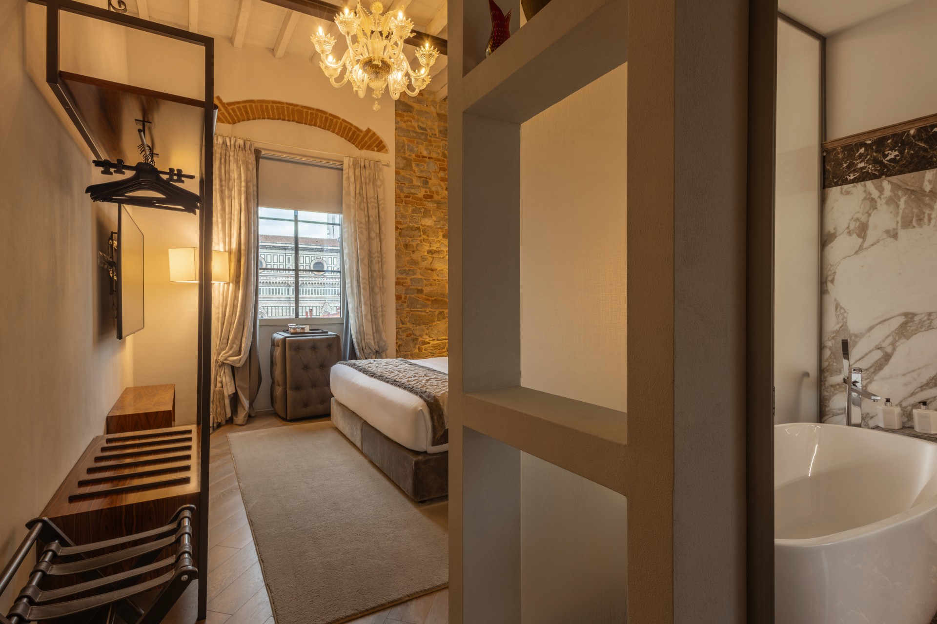 Modern hotel suite bedroom and bathroom entry with an open closet rack, double bed, exposed stone wall, crystal chandelier, and a soaking bathtub behind a partial wall.