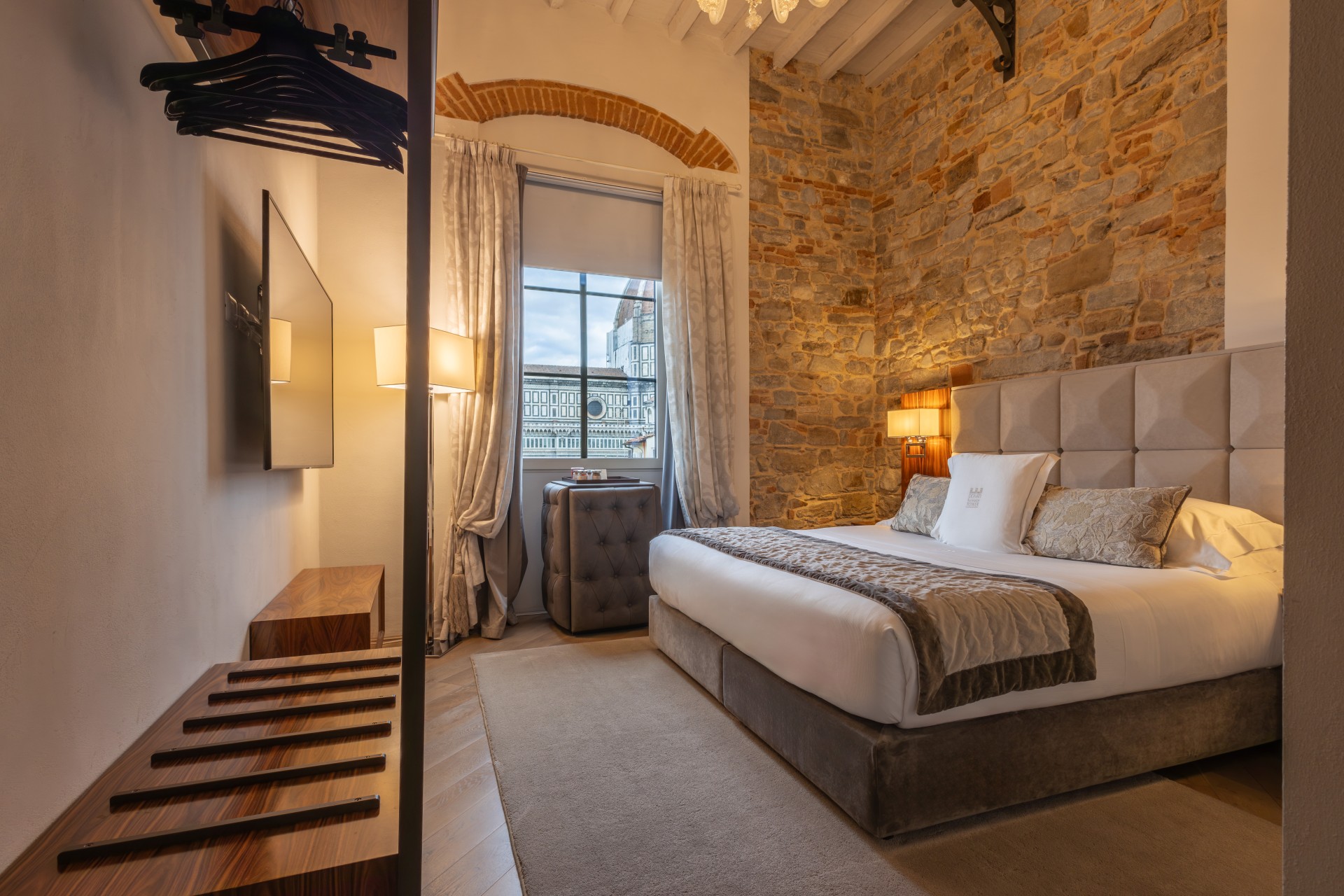Warmly lit hotel bedroom with a large upholstered bed, stone feature wall, open closet with luggage bench, wall-mounted TV, and window view of Florence Cathedral.