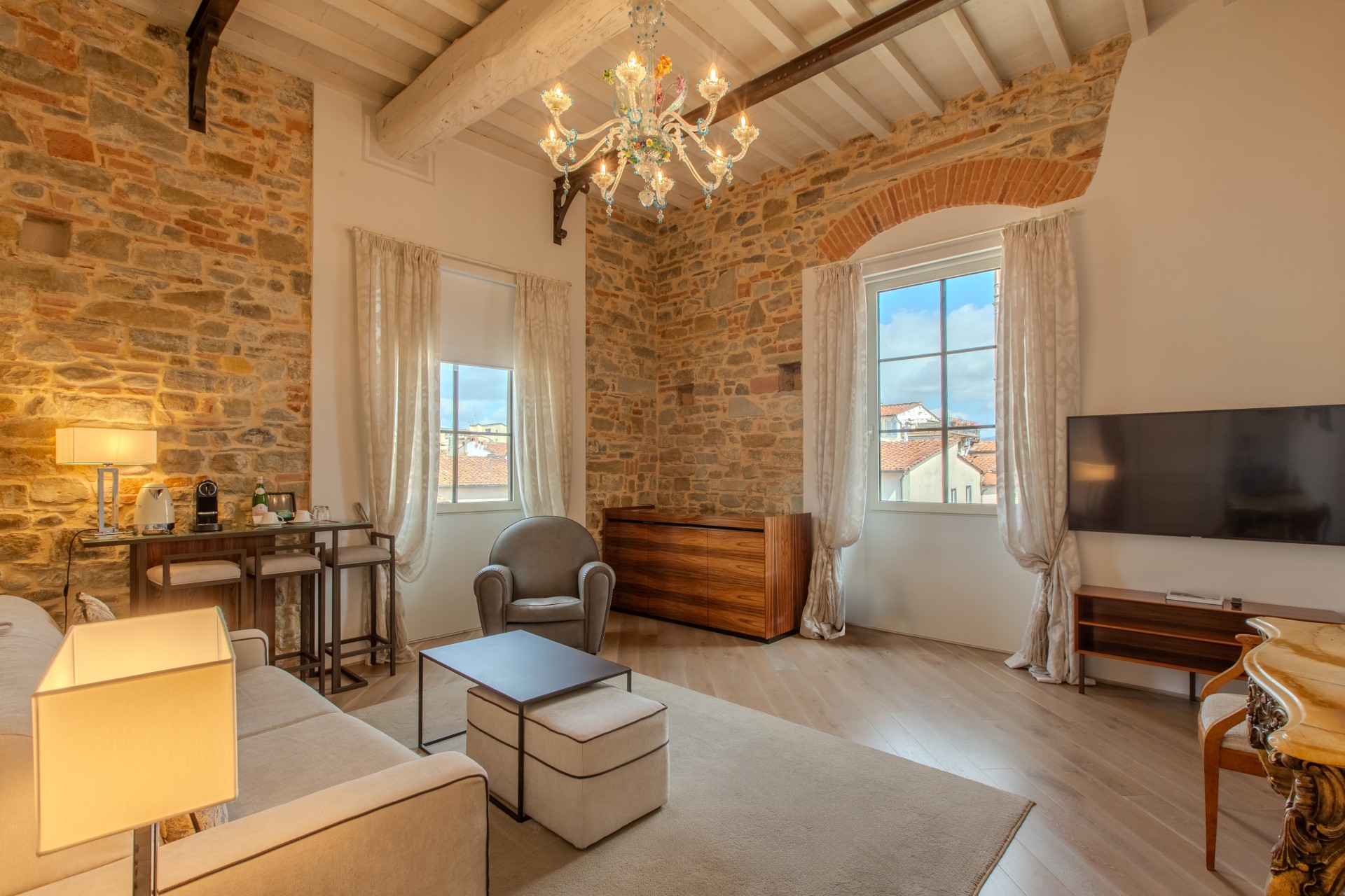 Spacious luxury suite living room with high beamed ceilings, glass chandelier, exposed stone walls, grey armchair, sofa, small bar area, and large windows.