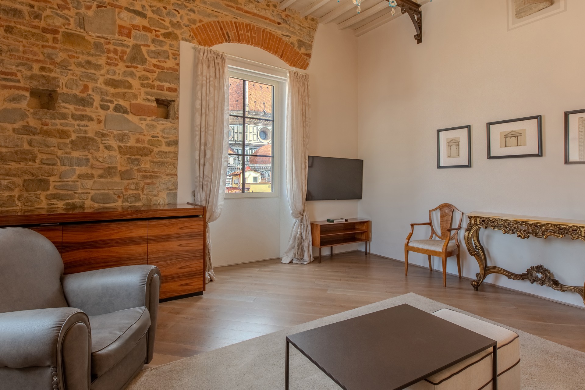 Elegantly styled hotel living room with exposed stone wall, wooden console table, grey armchair, low coffee table, and a window with a view of Florence Cathedral.