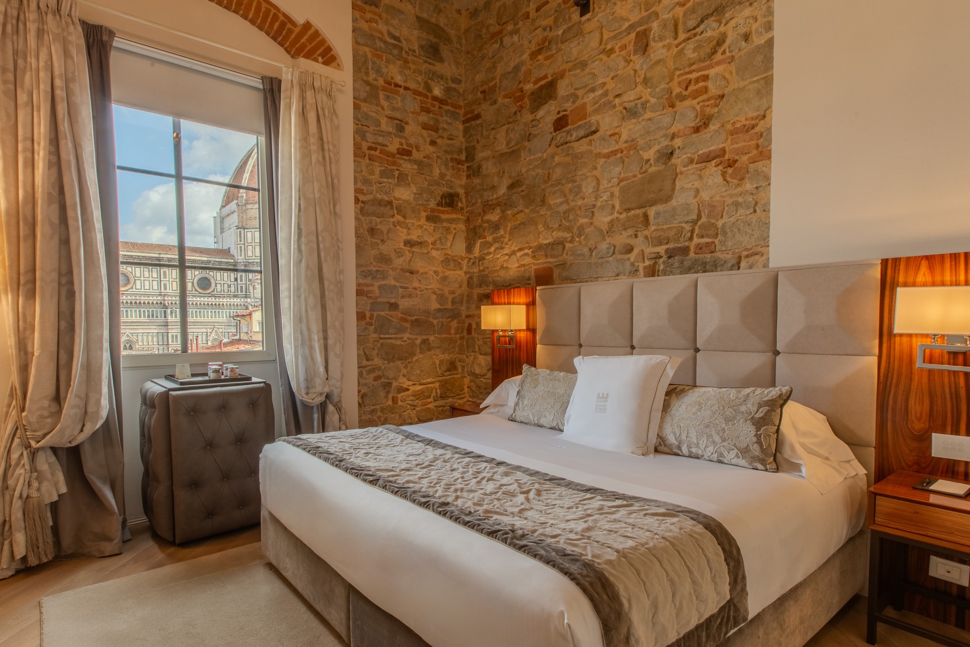 Hotel bedroom featuring a large upholstered bed, stone feature wall, warm bedside sconces, and a window with a close-up view of the Florence Cathedral dome.