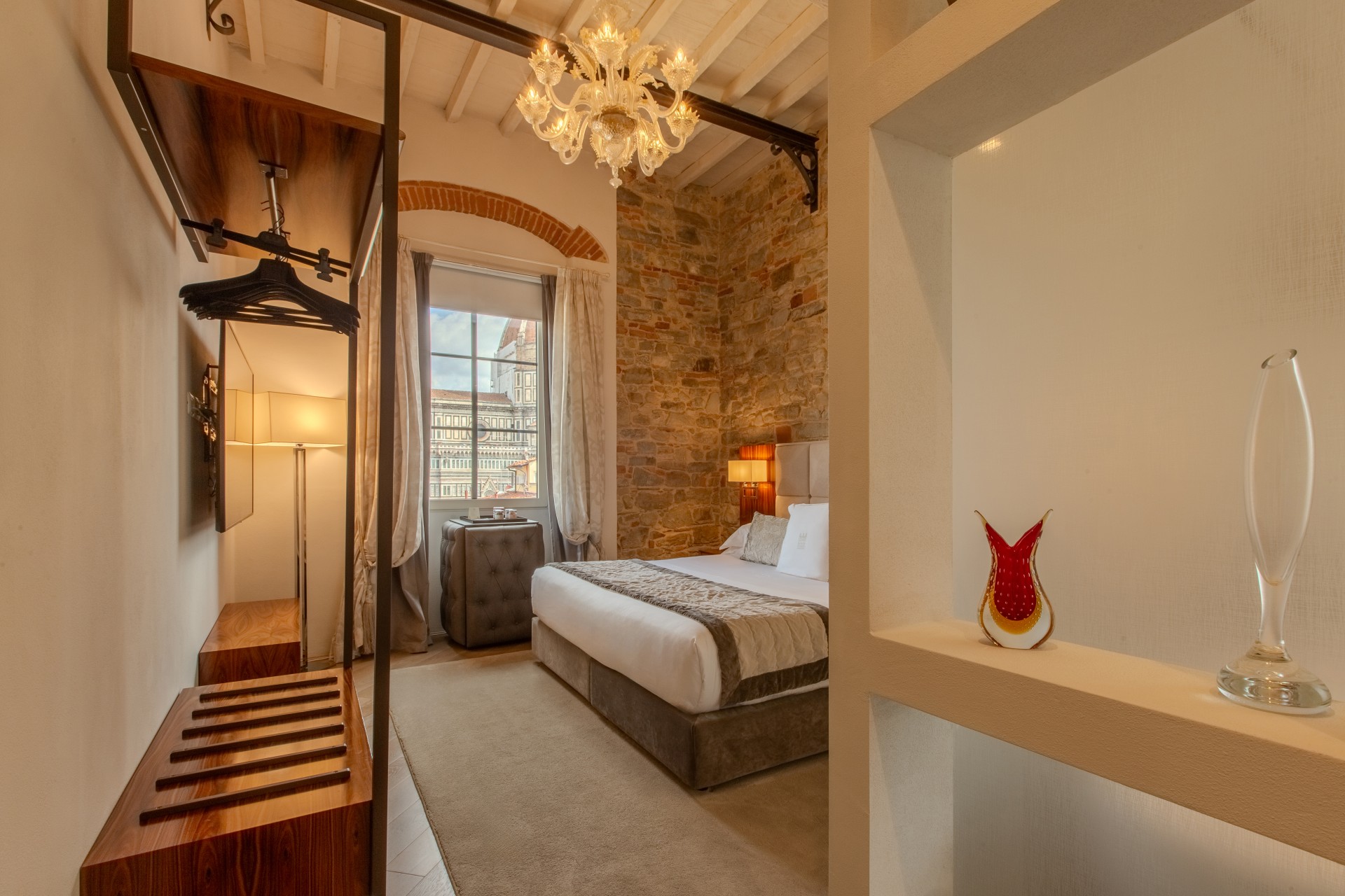 Hotel suite interior viewed past decorative glass vases, showing an open closet area, chandelier, king bed, stone wall, and window view of Florence Cathedral.