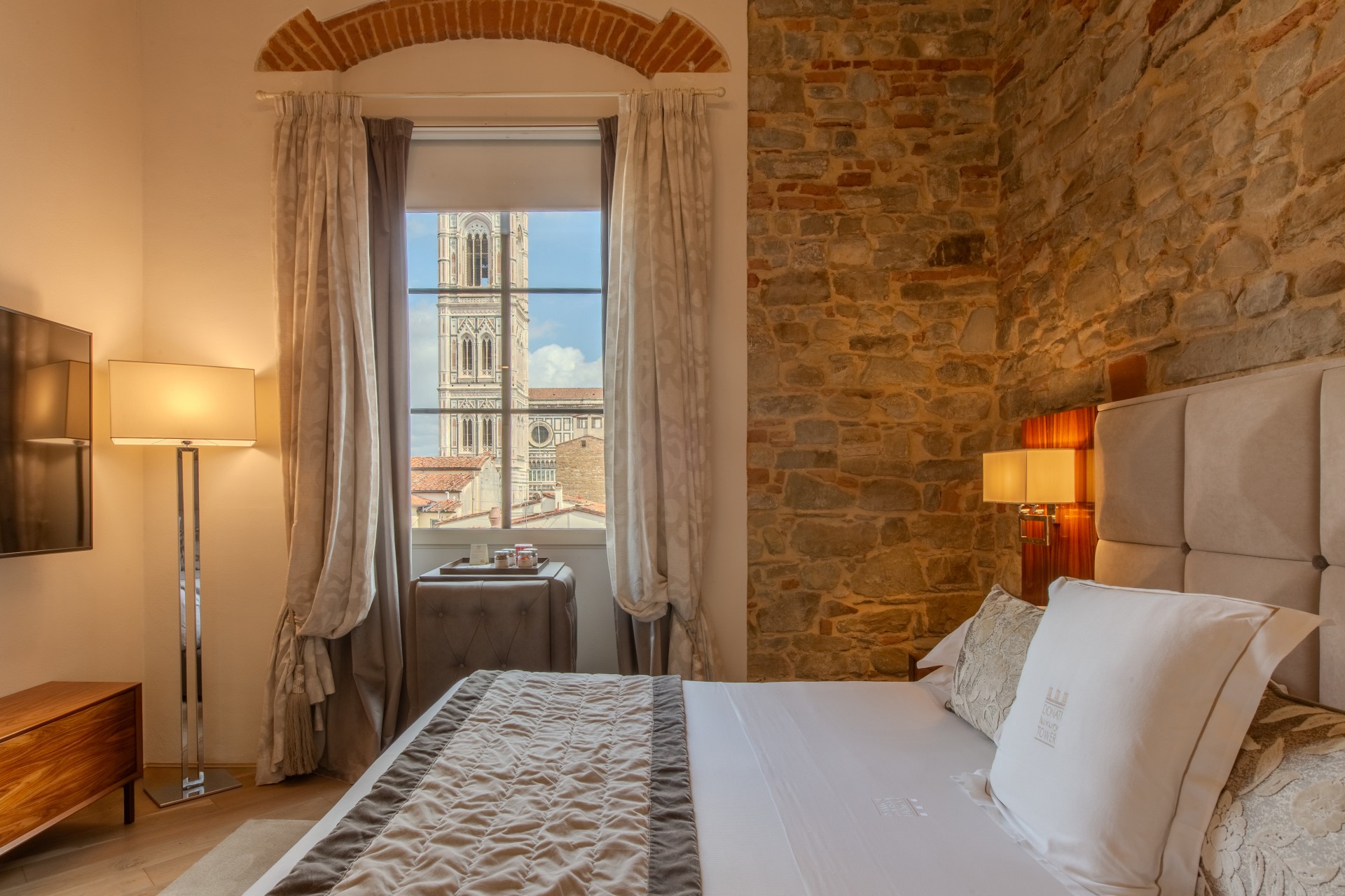 Hotel bedroom featuring exposed brick and stone walls, a plush headboard, soft floor lamp, and a window framing Giotto's Bell Tower in Florence.