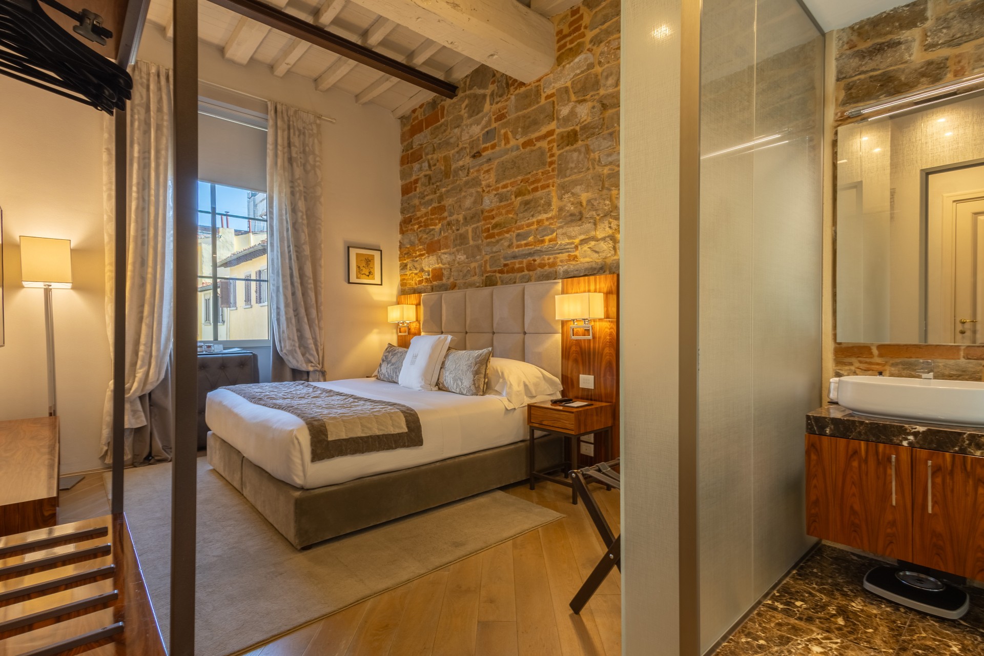 Hotel suite bedroom with an upholstered bed, exposed stone and brick accent wall, wooden ceiling beams, and an adjacent bathroom vanity visible through a frosted glass partition.