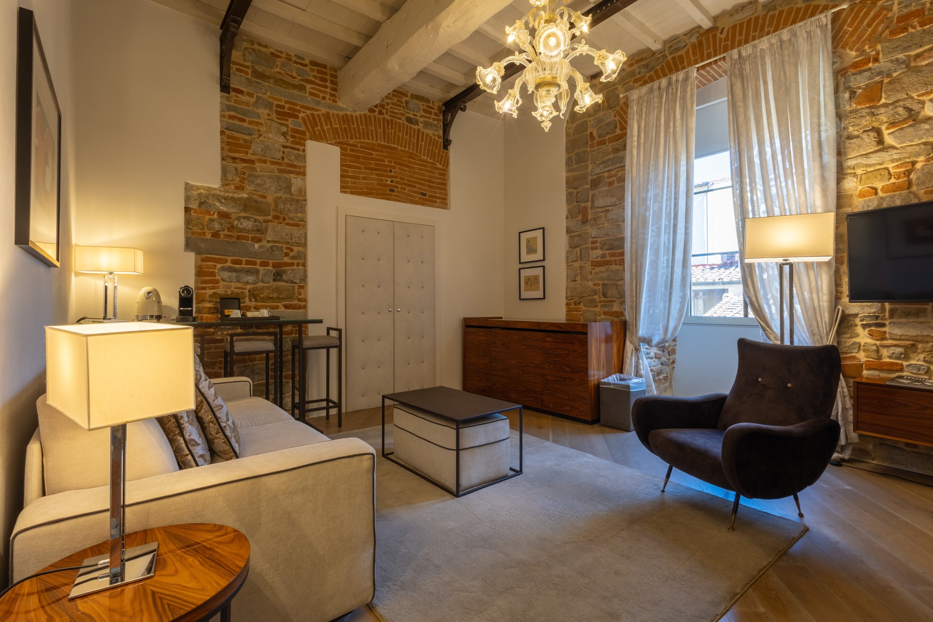 Spacious hotel suite living room with high beamed ceilings, exposed stone and brick accent walls, a grand glass chandelier, a beige sofa, a dark velvet armchair, and tall windows framed by sheer white curtains.