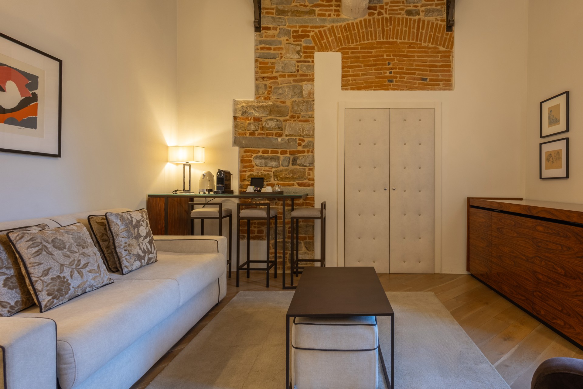 Hotel suite living room featuring a beige sofa with floral pillows, a low coffee table with a cushioned ottoman, a wood console cabinet, and a high counter table with bar stools set against an exposed brick wall.