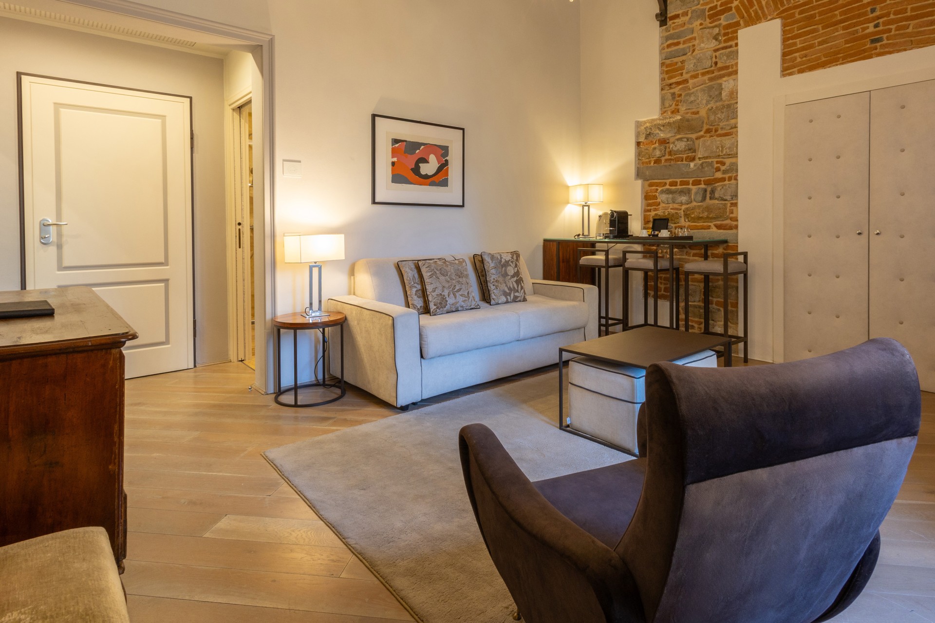 Living room in a hotel suite featuring light wood floors, a beige sofa, a dark armchair in the foreground, and an exposed brick and stone wall section near a mini-bar area.