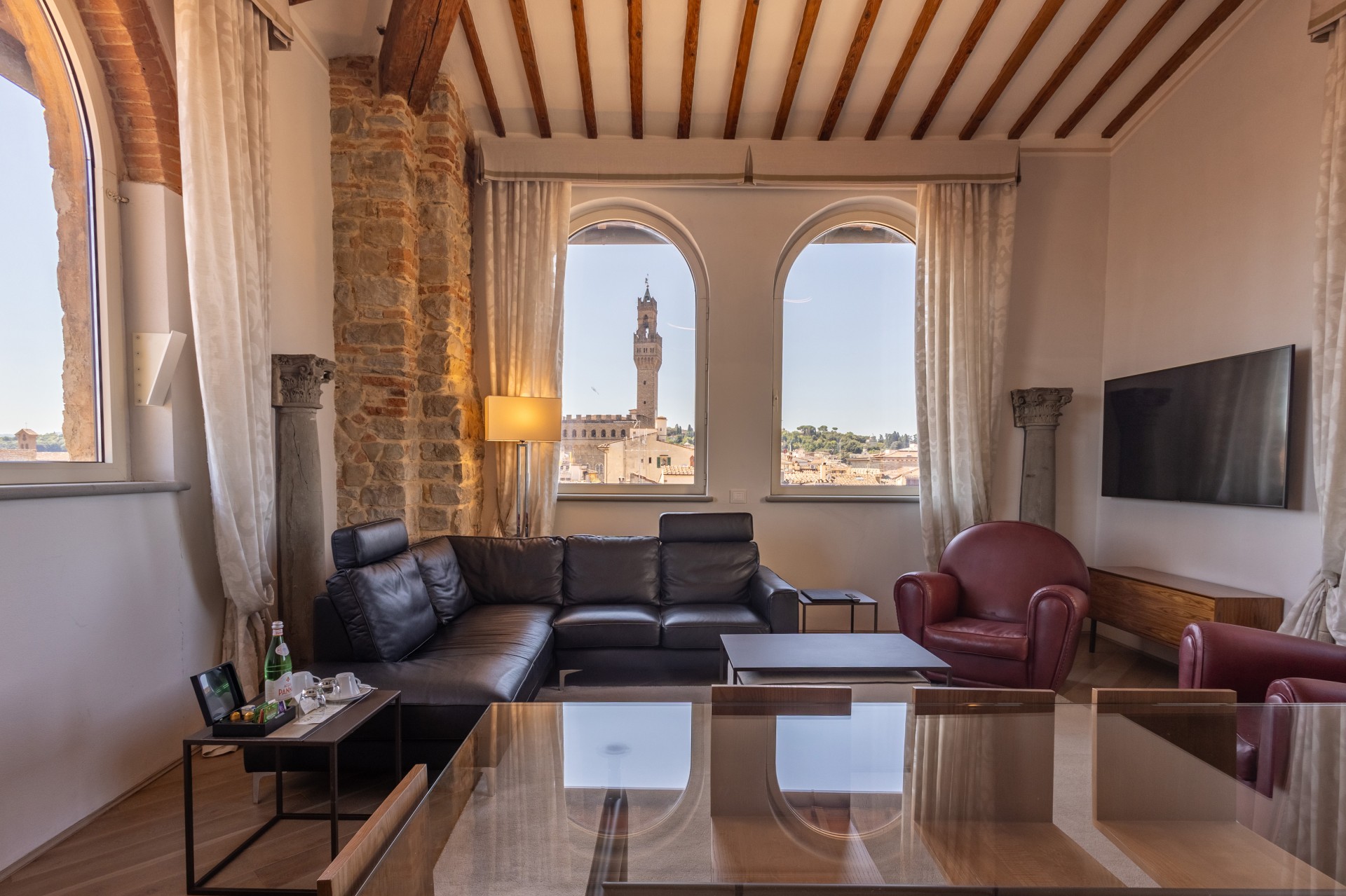 Luxurious suite living room featuring a black sectional sofa, red leather armchairs, exposed stone wall, ceiling beams, and arched windows framing a view of Palazzo Vecchio.