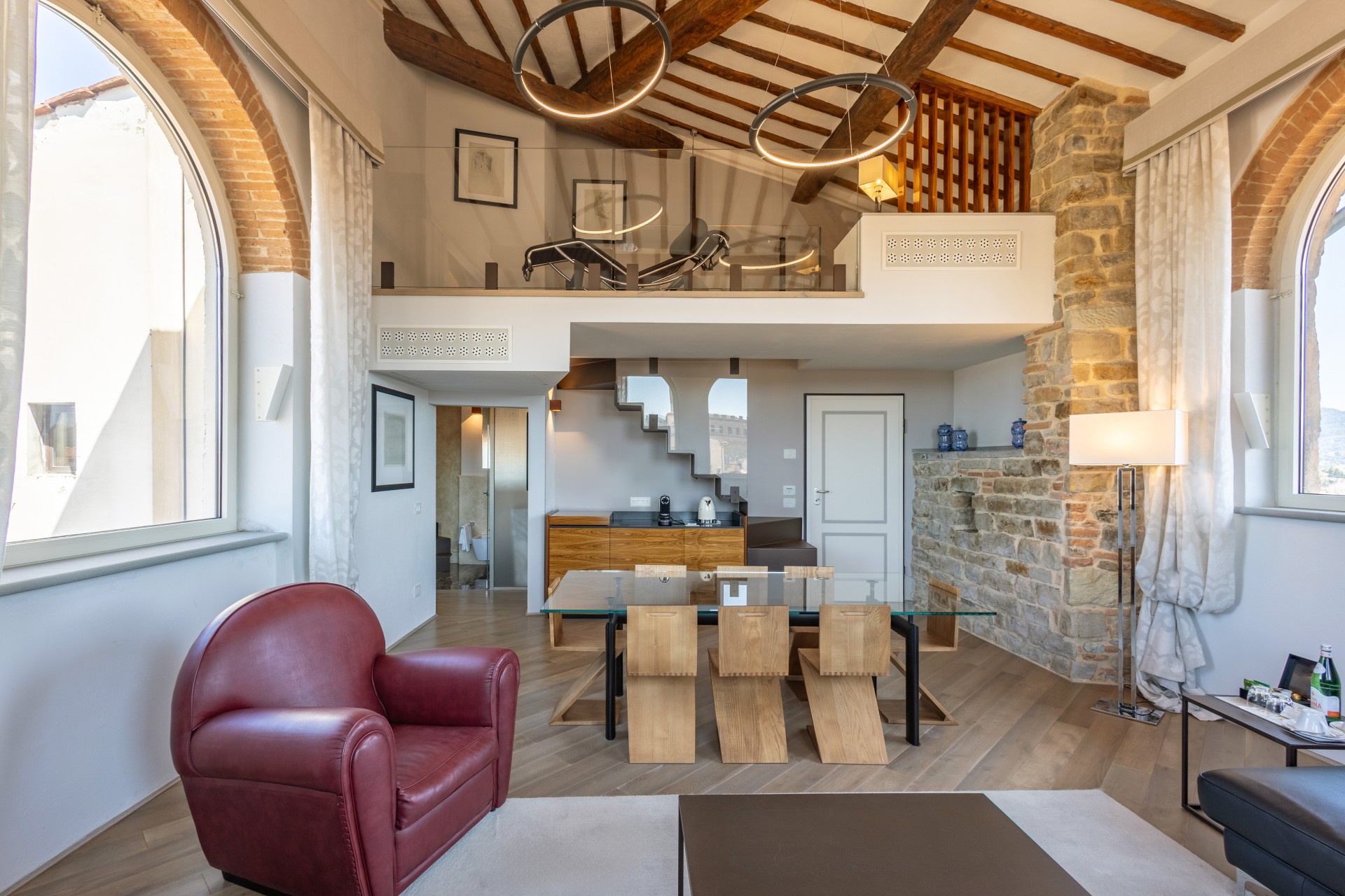 Modern multi-level suite featuring high vaulted ceilings with exposed wood beams, large arched windows, a glass-top dining table with wooden chairs, a red leather armchair, and a loft mezzanine level.