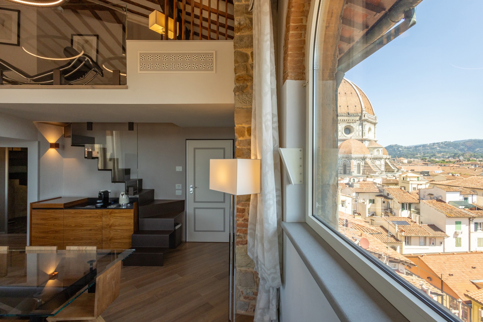 Modern multi-level suite in Florence featuring a large arched window with a direct view of the Florence Cathedral (Duomo), a floating staircase leading to a mezzanine, wooden ceilings, and a kitchenette.