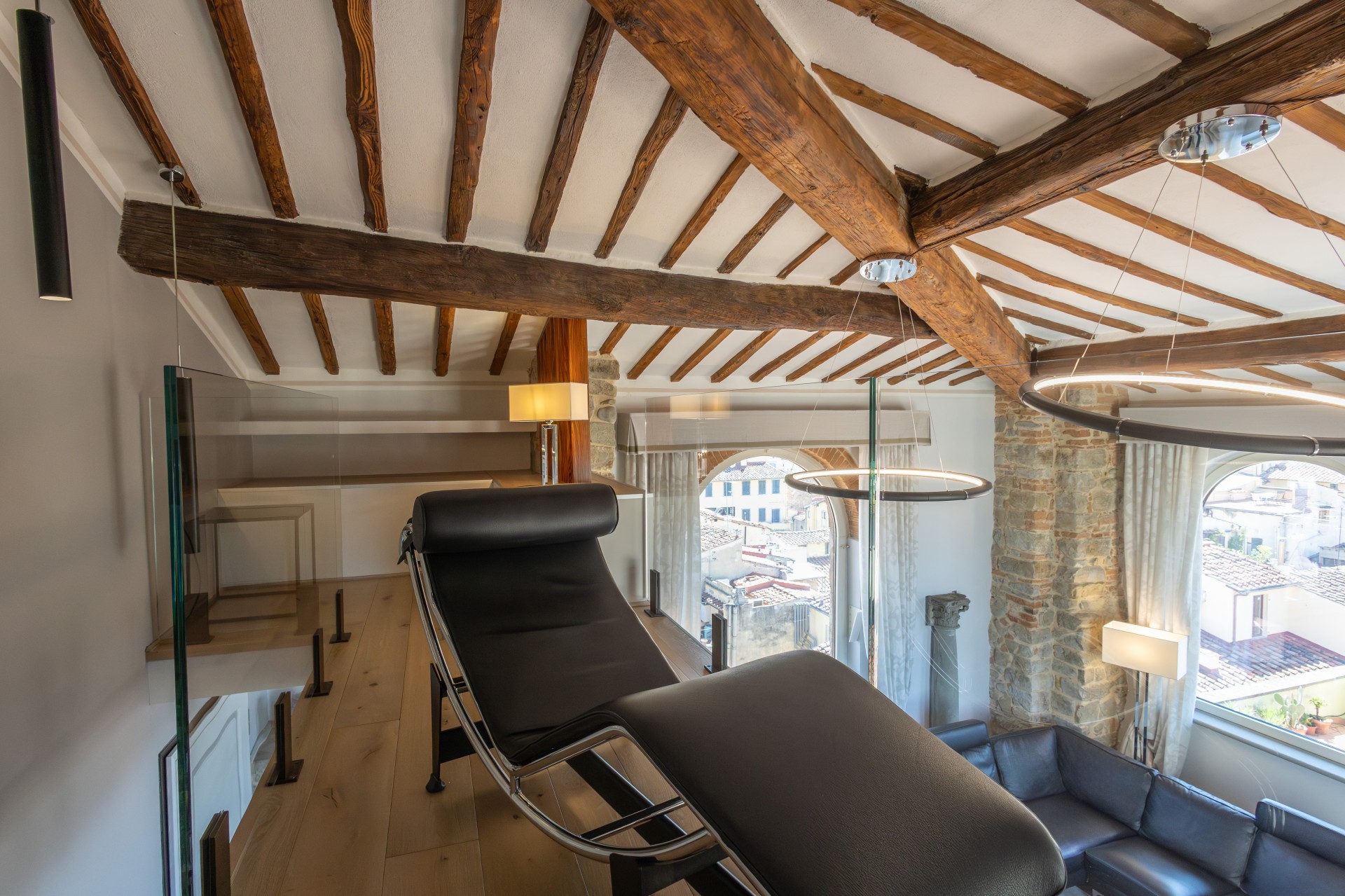 Mezzanine level in a luxury suite featuring a black leather chaise lounge, glass railings, exposed wooden ceiling beams, modern ring pendant lights, and large windows overlooking city rooftops.