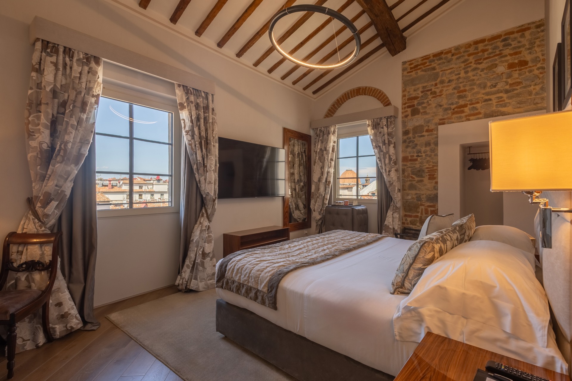 Elegant hotel bedroom featuring a large double bed, exposed brick wall, vaulted ceiling with wooden beams, modern ring light fixture, wall-mounted flat-screen TV, and windows with city views.