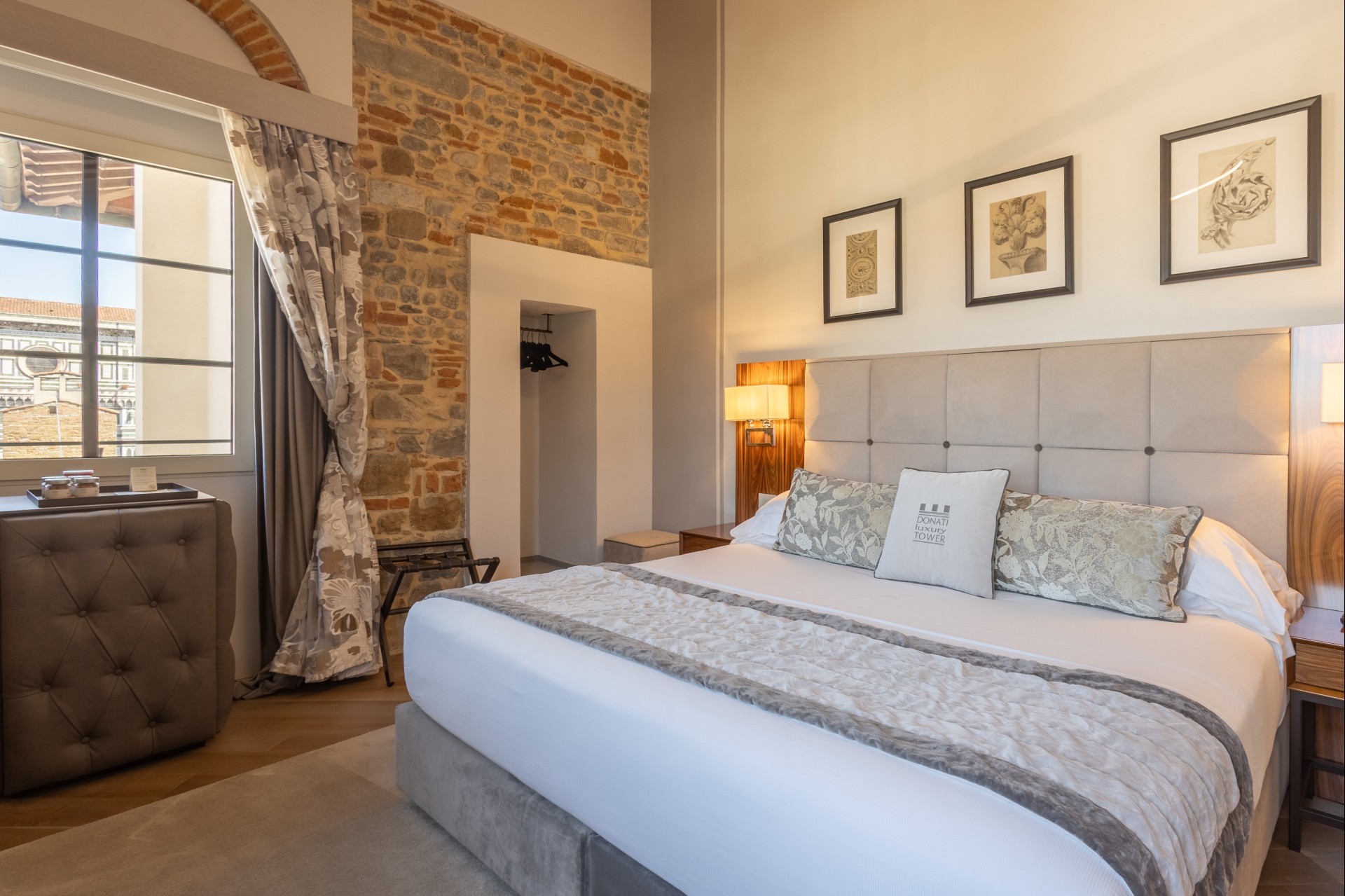 Hotel bedroom featuring a large upholstered bed with decorative pillows, framed wall art, exposed stone brick wall, tufted bar cabinet, and a large window with patterned drapes.