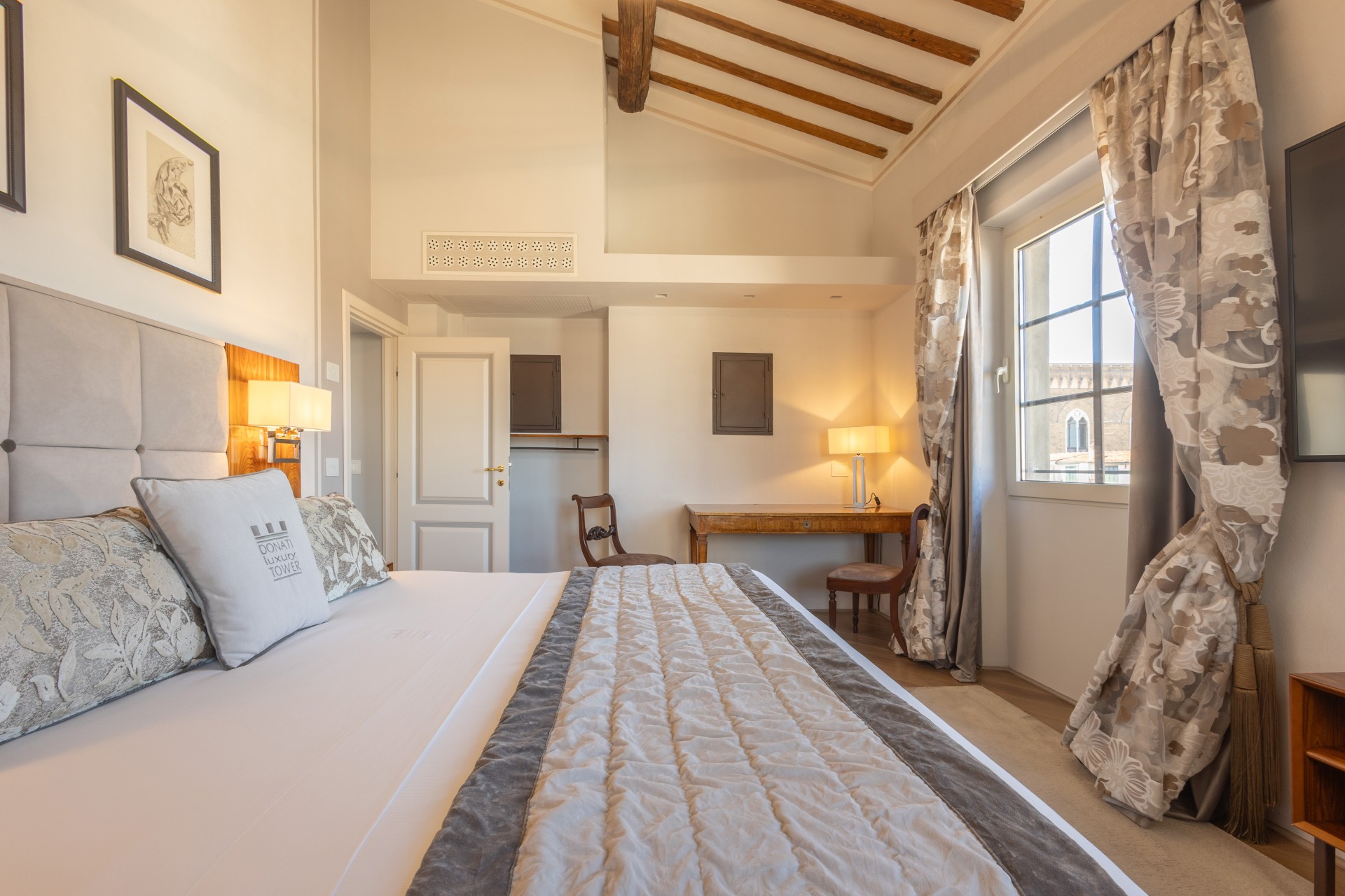 Bright hotel bedroom featuring a plush bed with decorative pillows, a wooden writing desk, high sloped ceilings with wooden beams, framed artwork, and patterned floor-length drapes beside a window.