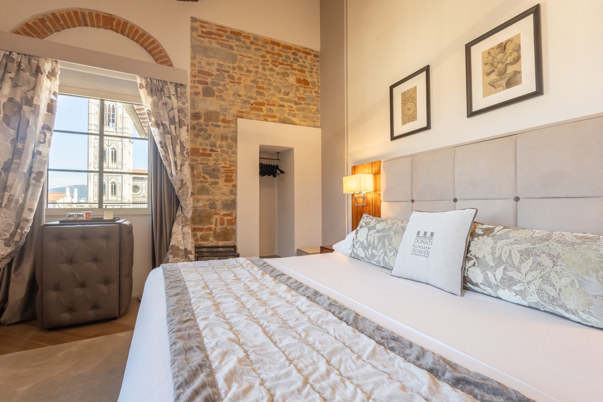 Spacious hotel bedroom with a plush double bed, exposed stone and brick accent wall, framed art, and a large window offering a view of Giotto's Bell Tower.