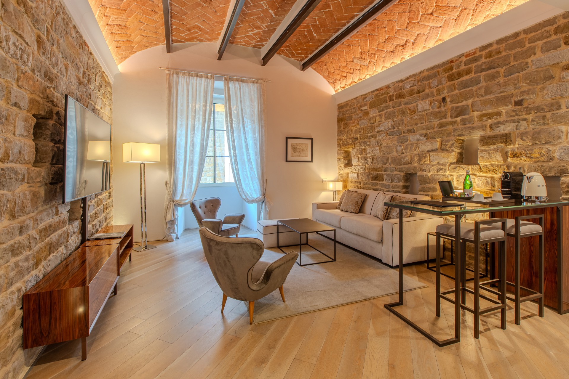 A wide angle view of a luxury hotel suite living room with exposed stone walls, a vaulted terracotta brick ceiling with wooden beams, a wall-mounted TV, plush armchairs, sofa, and a glass-topped high-top bar table.