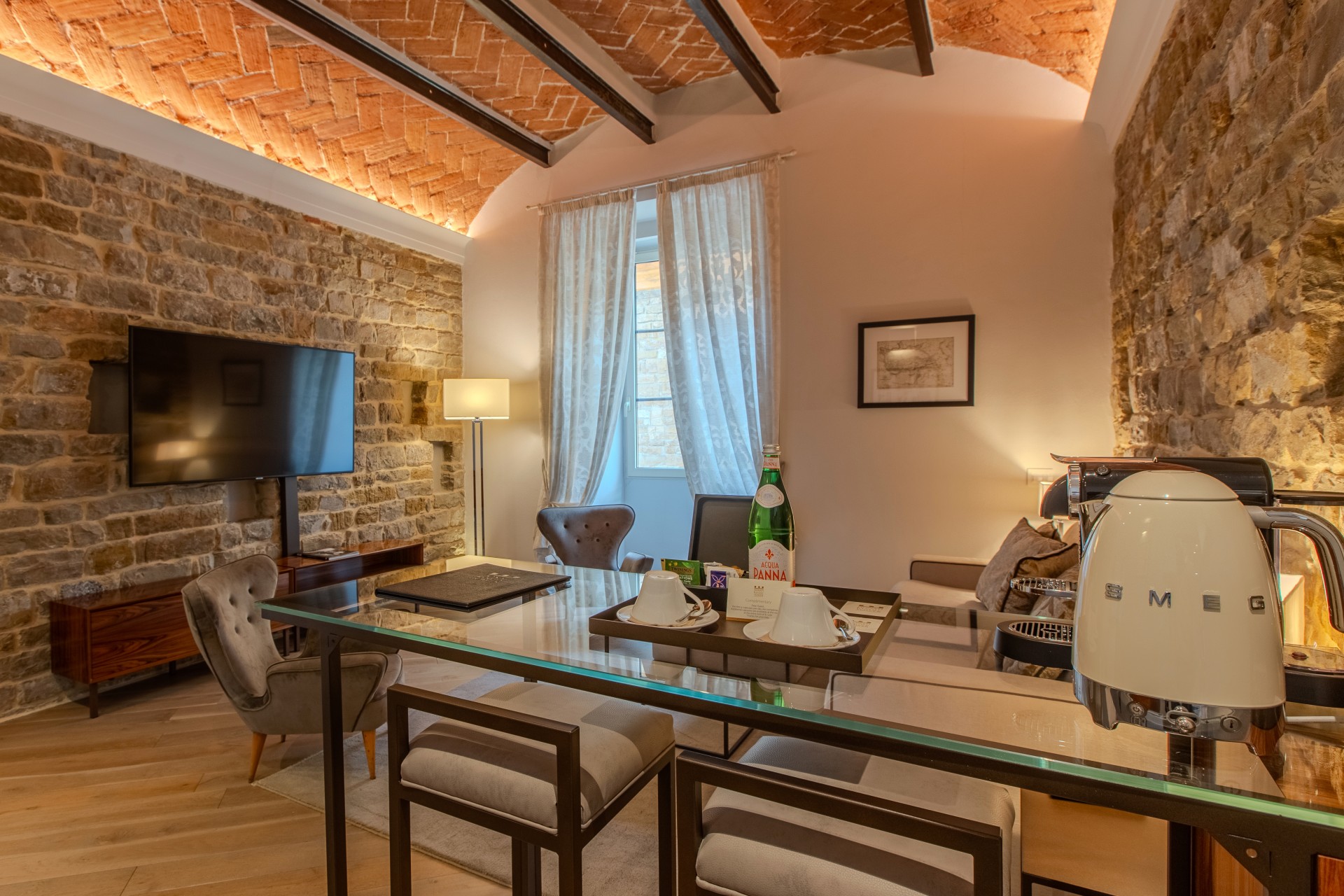 A luxury hotel suite featuring exposed stone walls, a vaulted brick ceiling, a glass-topped high-top table set with teacups, a Smeg kettle, and bar stools, leading into a cozy seating area with a wall-mounted TV.
