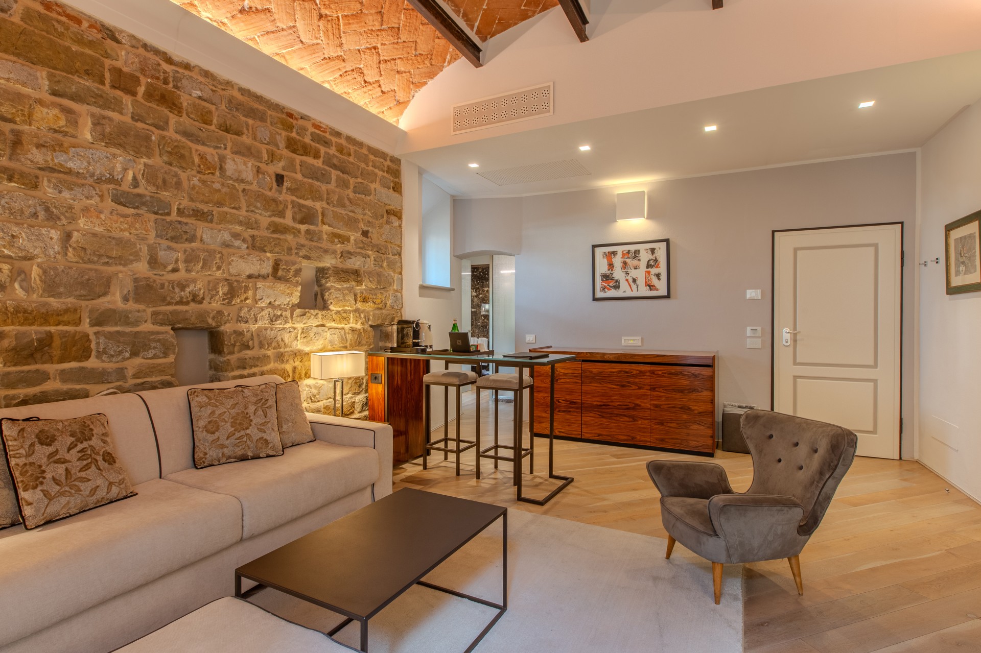 A luxury hotel suite living room showcasing exposed stone walling, a vaulted herringbone brick ceiling with warm lighting, a cream couch with accent pillows, a grey velvet armchair, a coffee table, and a raised breakfast bar with bar stools.