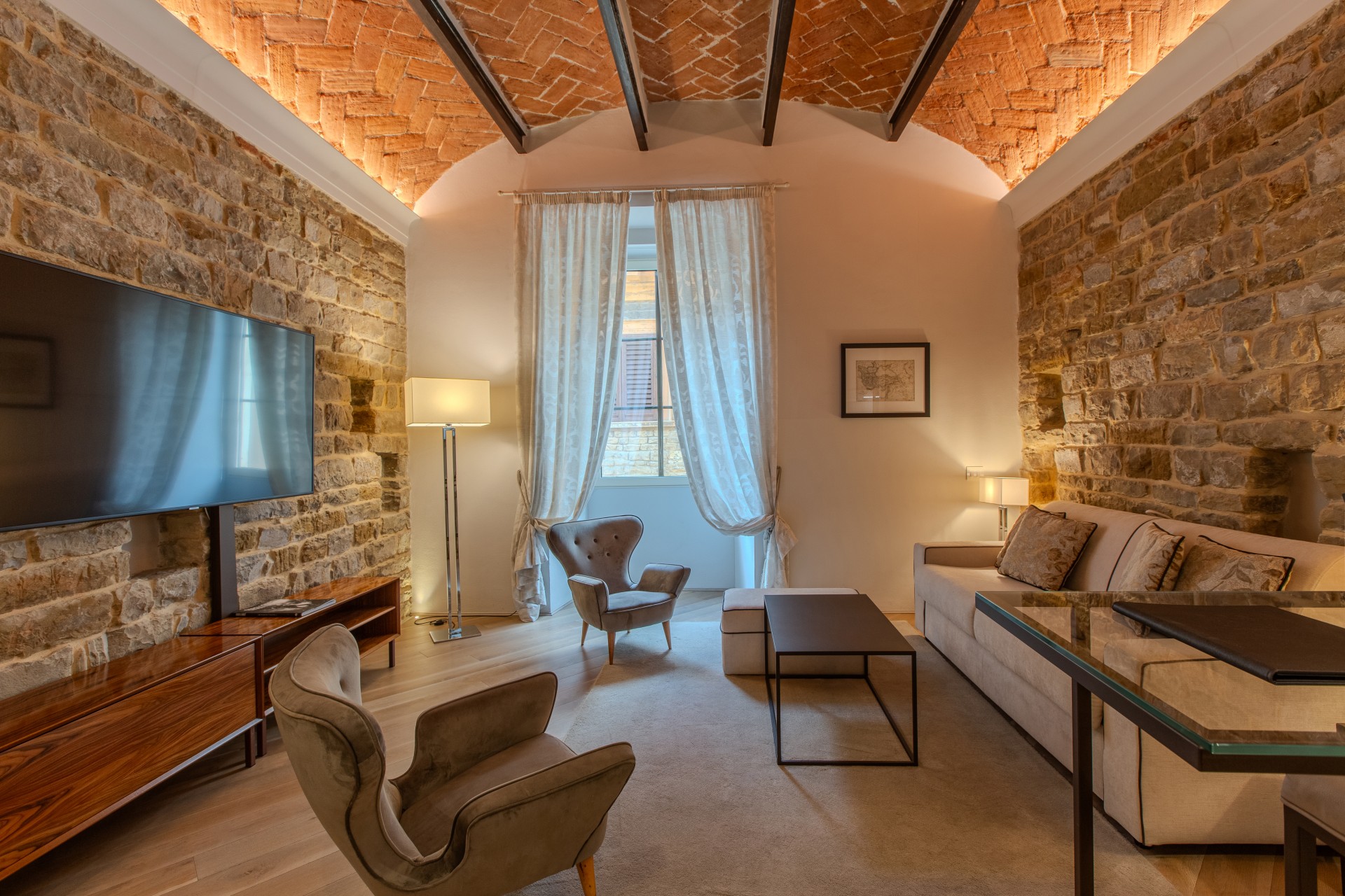 A luxury hotel suite living room featuring exposed stone walls, a vaulted brick ceiling with dark wooden beams, a large wall-mounted TV, beige sofa, velvet armchairs, and floor-to-ceiling sheer curtains.