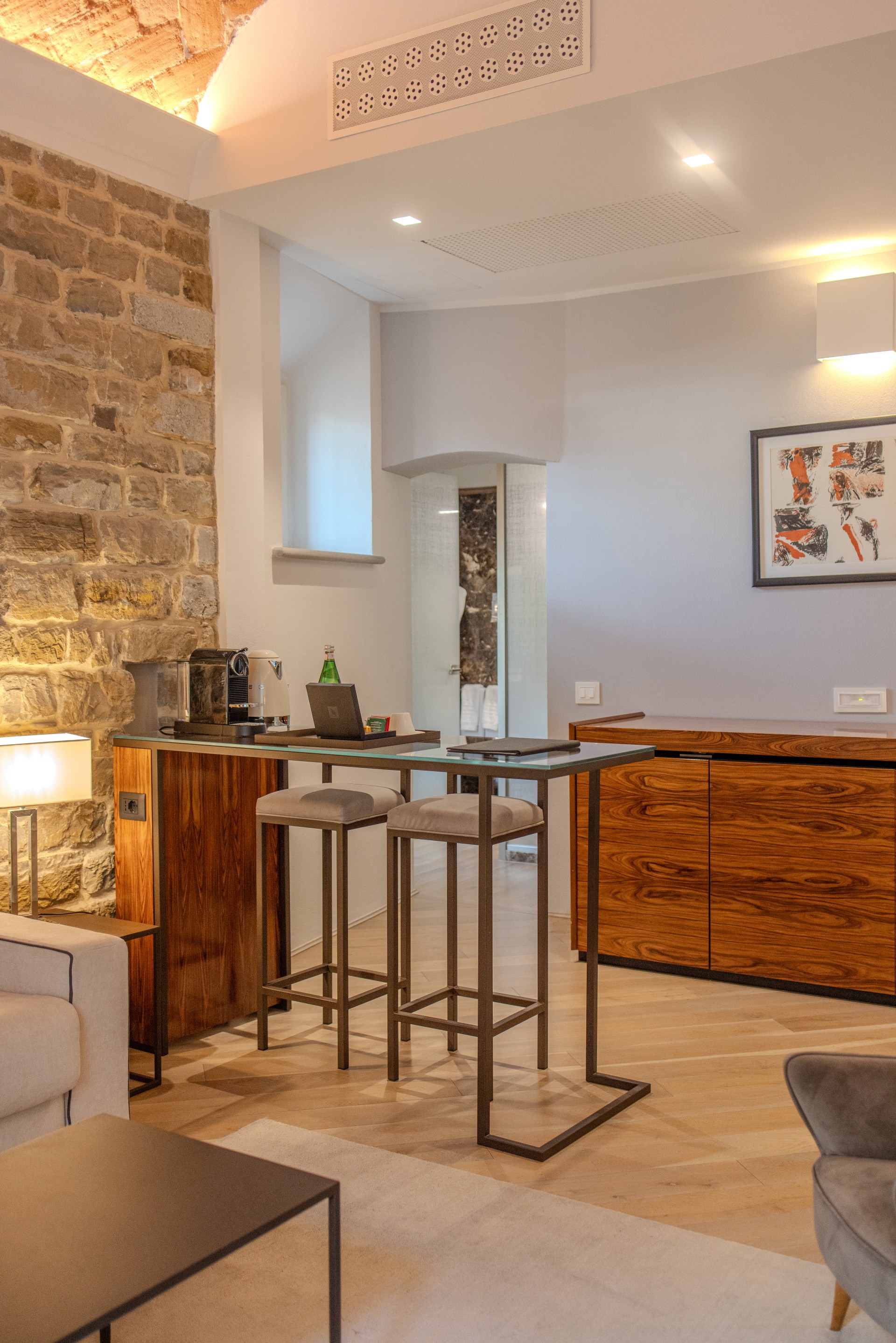 A modern hotel suite interior featuring an exposed stone feature wall, a glass-topped high-top table with two bar stools, a coffee station, a rich wood credenza, and light hardwood flooring.
