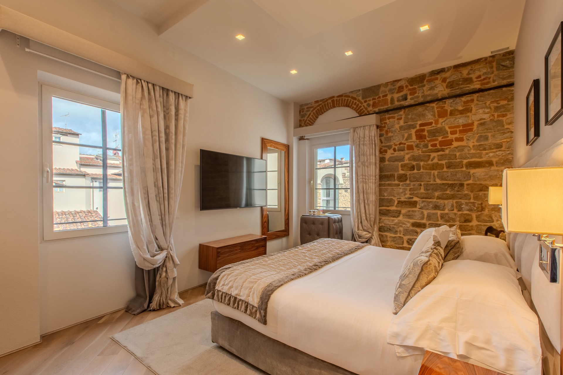 A cozy hotel bedroom with an exposed stone wall, a large bed with crisp white linens and a quilted runner, a wall-mounted flat-screen TV, wooden floating console, and bright windows offering views of European-style architecture.
