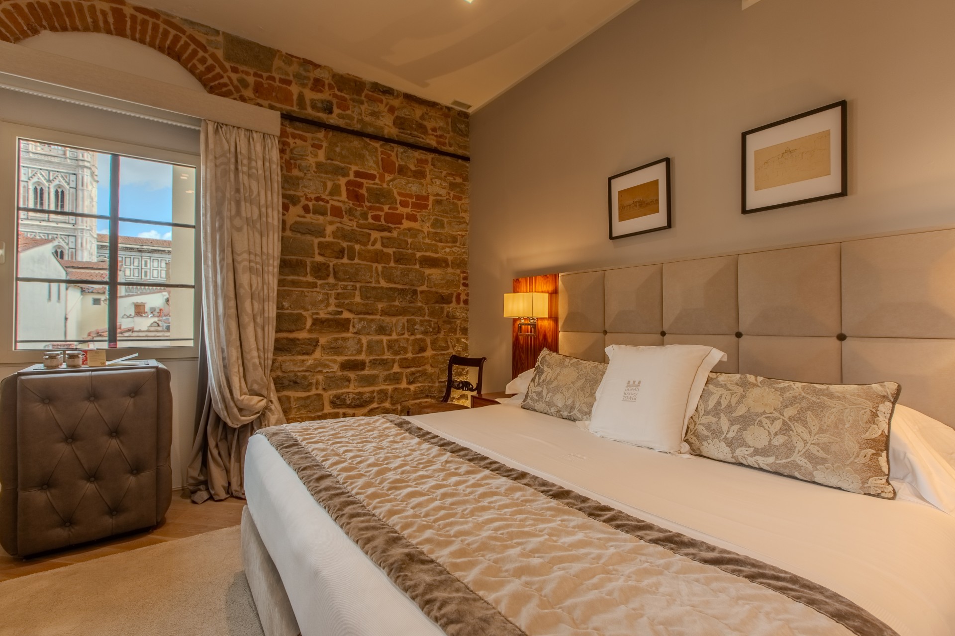 An elegant hotel bedroom featuring a beige upholstered bed, an exposed stone wall, framed artwork, and a window framing a view of Florence.