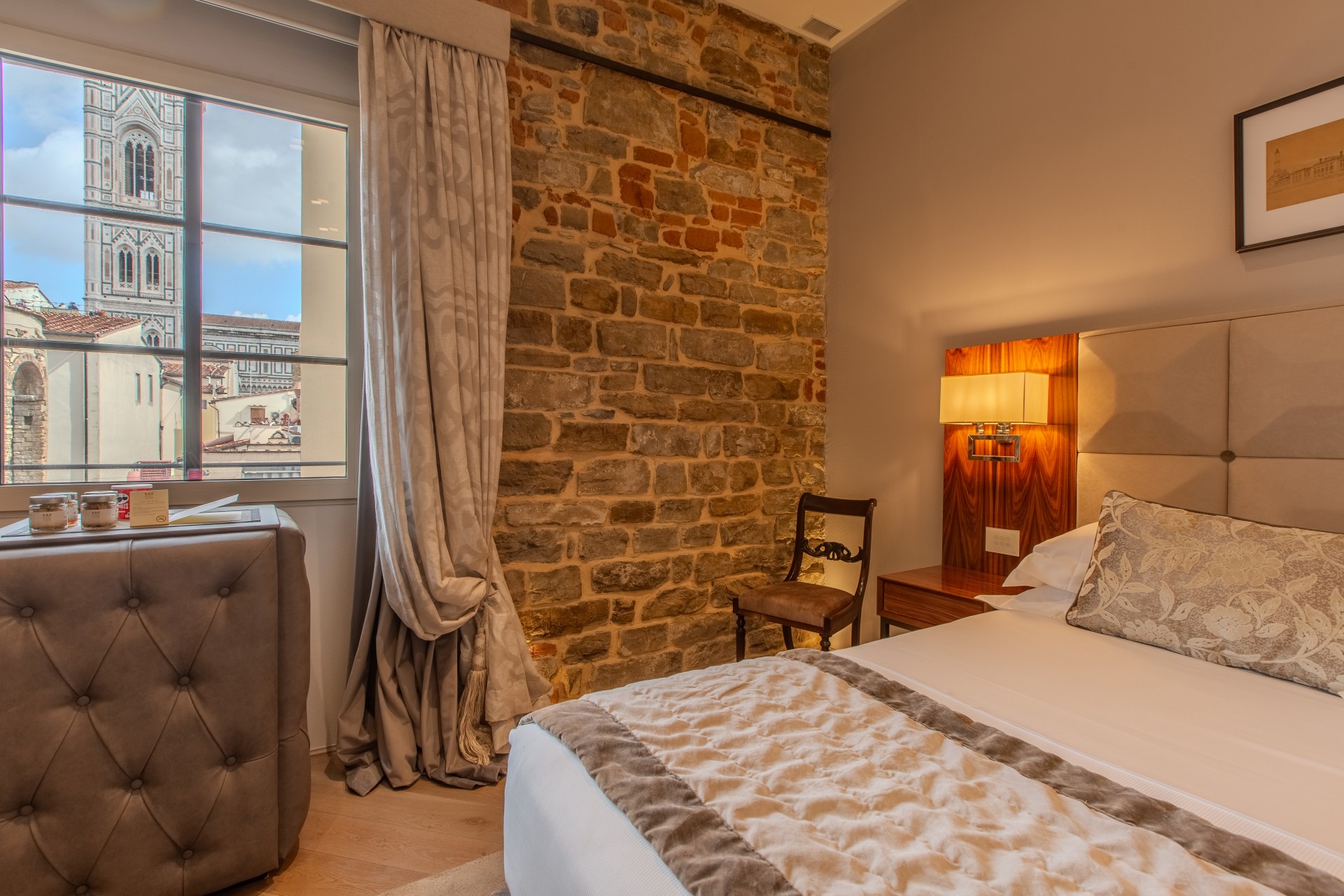 A cozy hotel bedroom with an exposed brick feature wall, a bed dressed in beige linens, and a window offering a view of Giotto's Campanile in Florence.