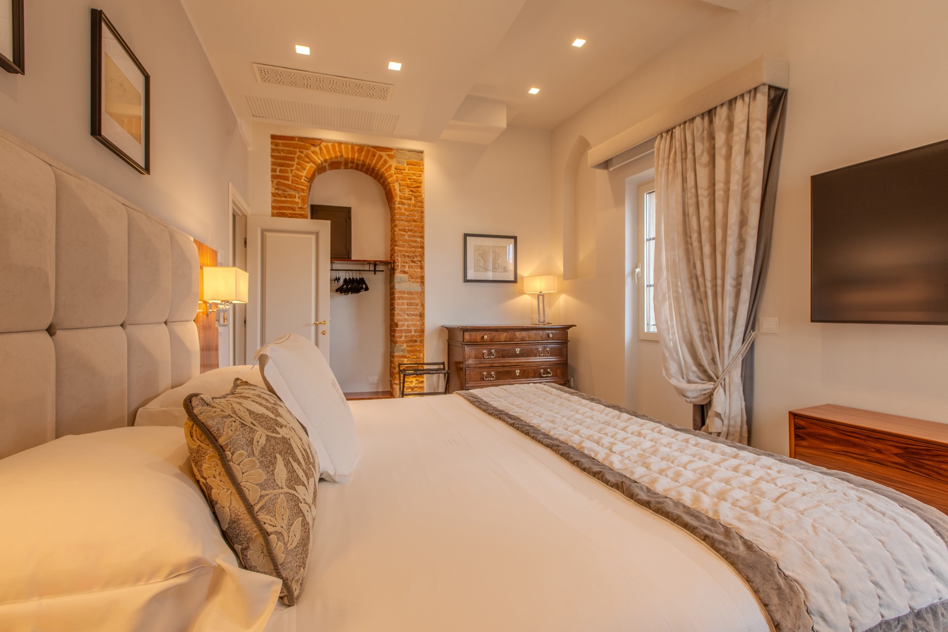 An elegant hotel bedroom featuring a plush king-size bed with a tufted headboard, an exposed brick arched nook, a wall-mounted TV, a dark wood dresser, and warm ambient lighting.
