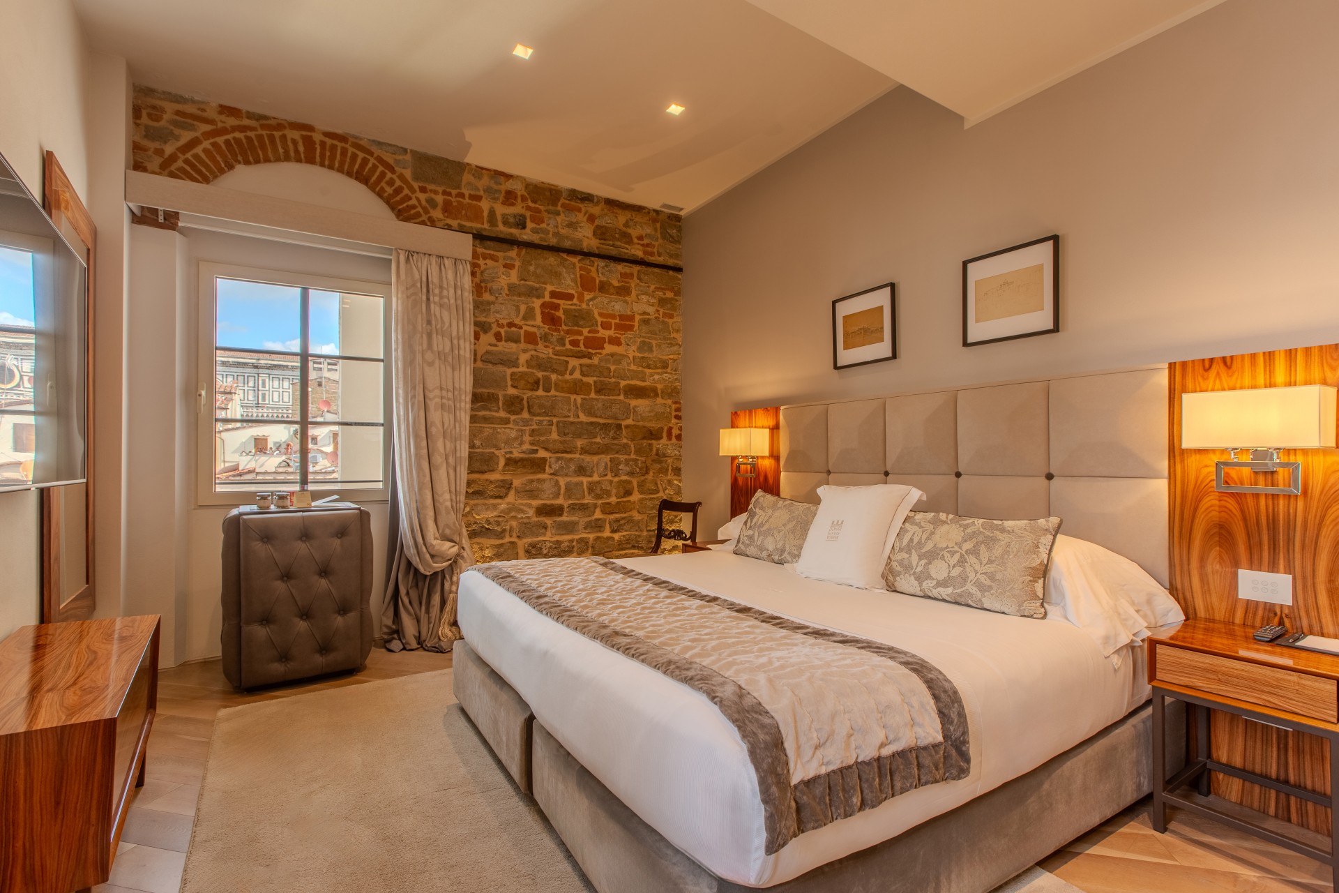 A stylish hotel bedroom with a plush king-size bed, upholstered tufted headboard, warm wood nightstands, an exposed stone wall feature, and a window overlooking historic architecture.