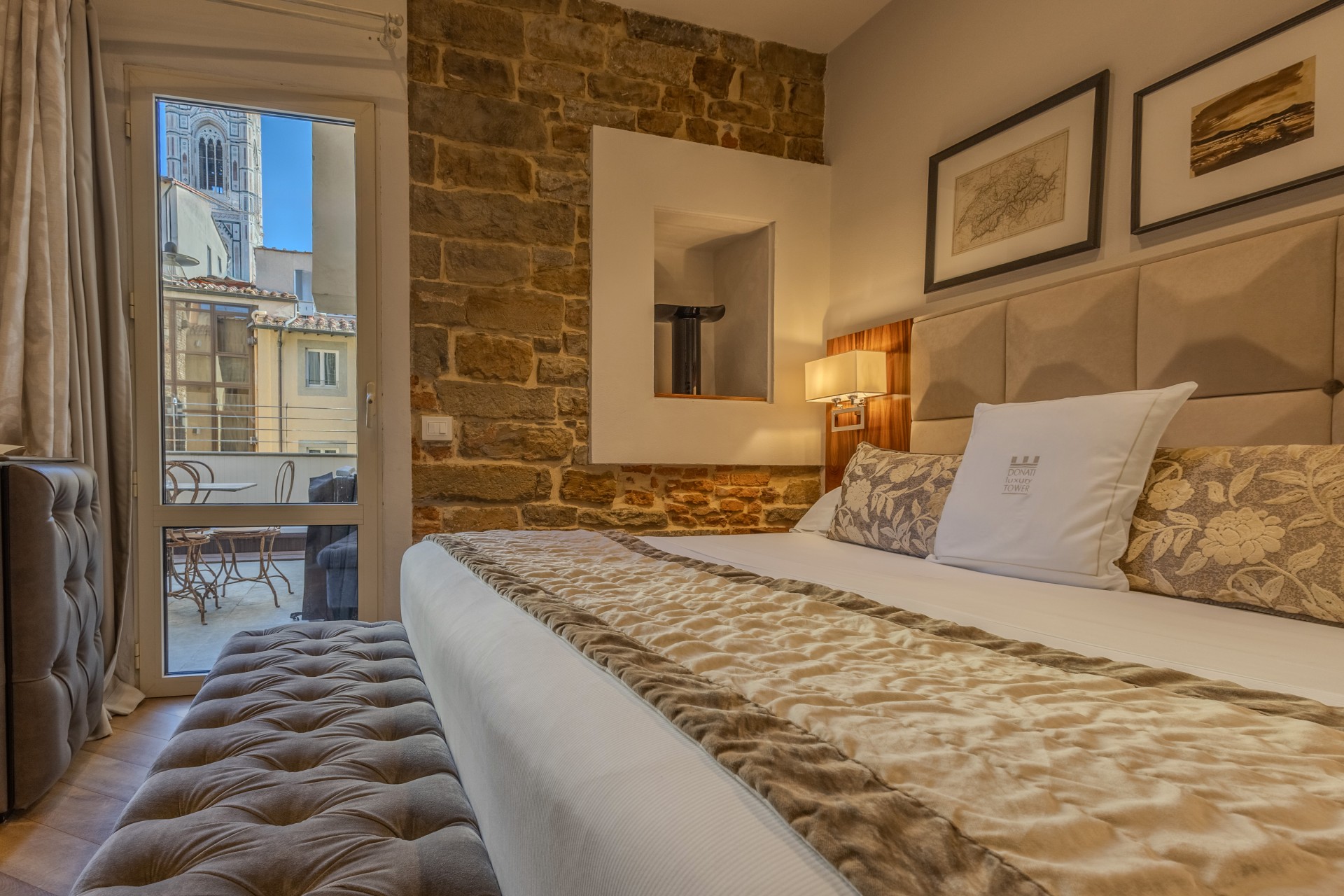 Cozy hotel room featuring a king-size bed with patterned pillows, an tufted ottoman bench, exposed stone brickwork, and a balcony doorway offering views of historic architecture.