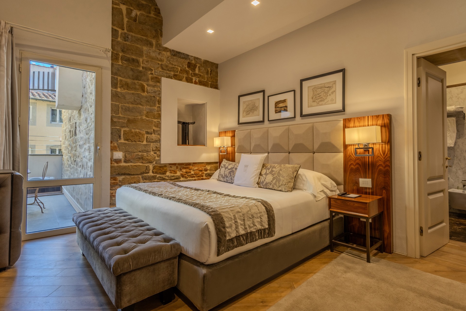 Warmly lit hotel room featuring a king-size bed with a tufted headboard and plush bench, an exposed stone wall, framed artwork, and glass doors opening to a private balcony.