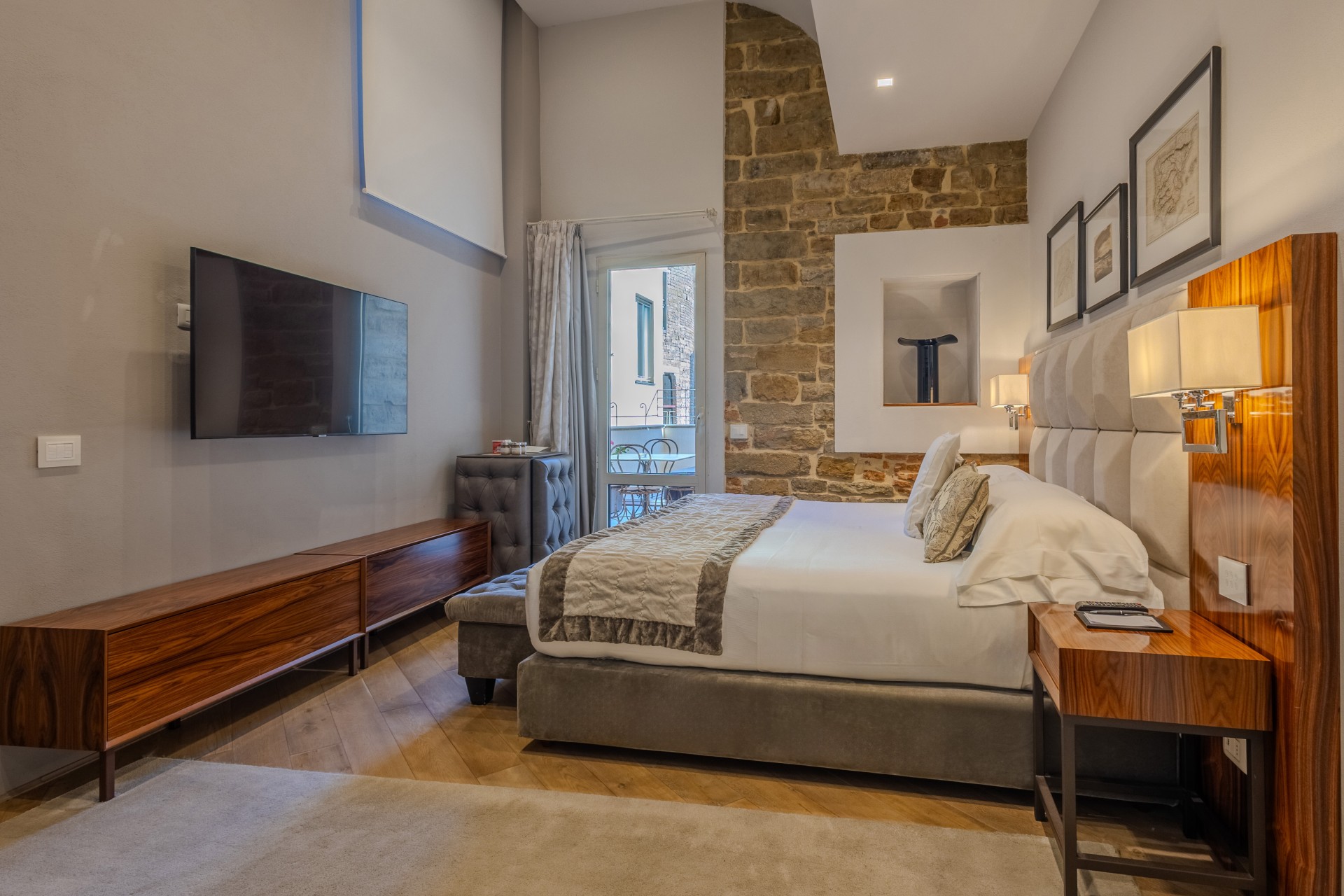 Modern hotel room with a flat-screen TV, a low wooden media console, a king-size bed, exposed stone accent wall, and a balcony door.