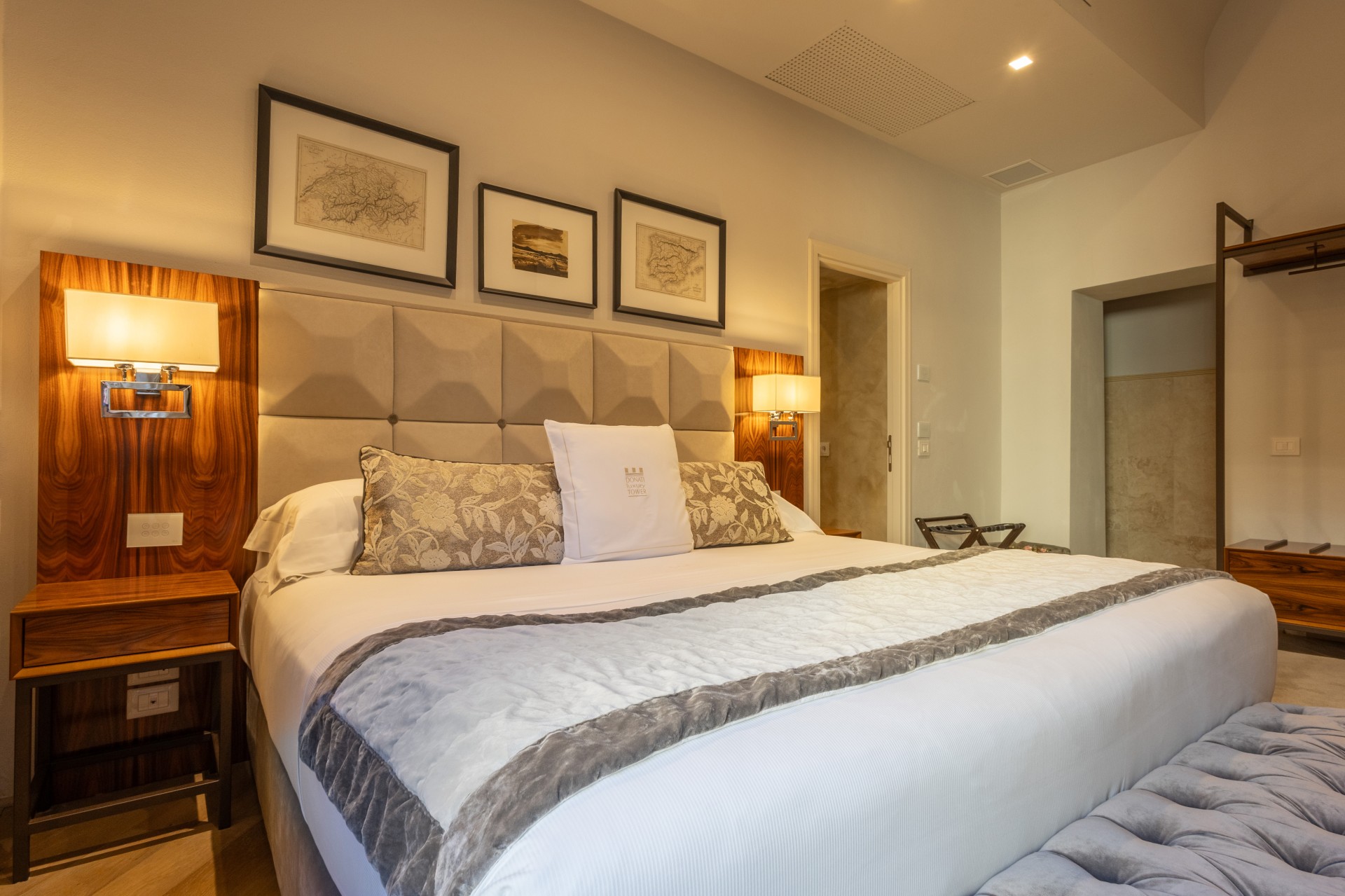 Hotel bedroom suite with a king-size bed, tufted beige headboard, warm wooden nightstands with mounted lamps, framed wall art, and an upholstered bench at the foot of the bed.