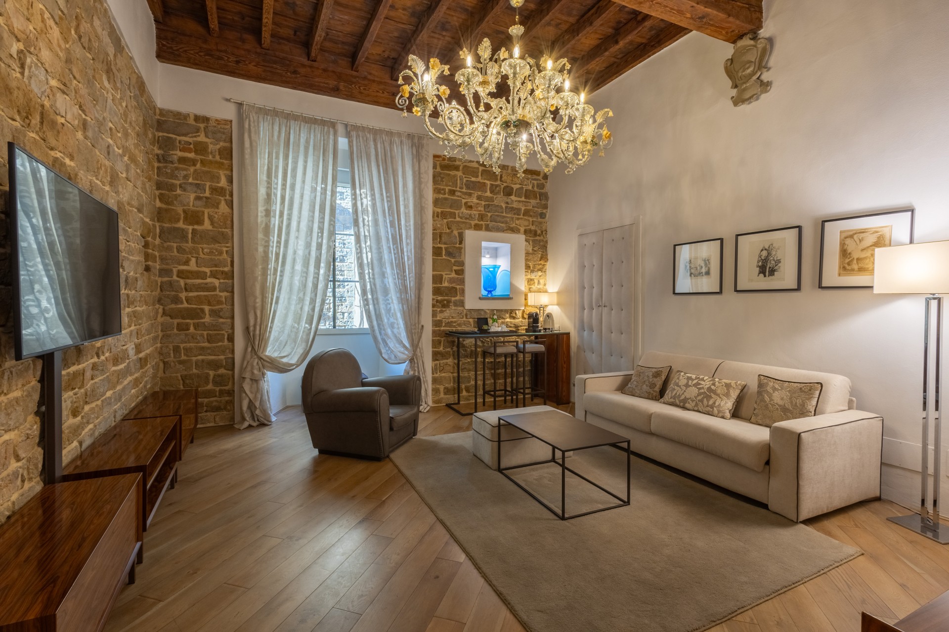 Spacious luxury suite living room with exposed stone, a large TV, wooden ceiling beams, an ornate crystal chandelier, and a beige sofa set over a neutral area rug.