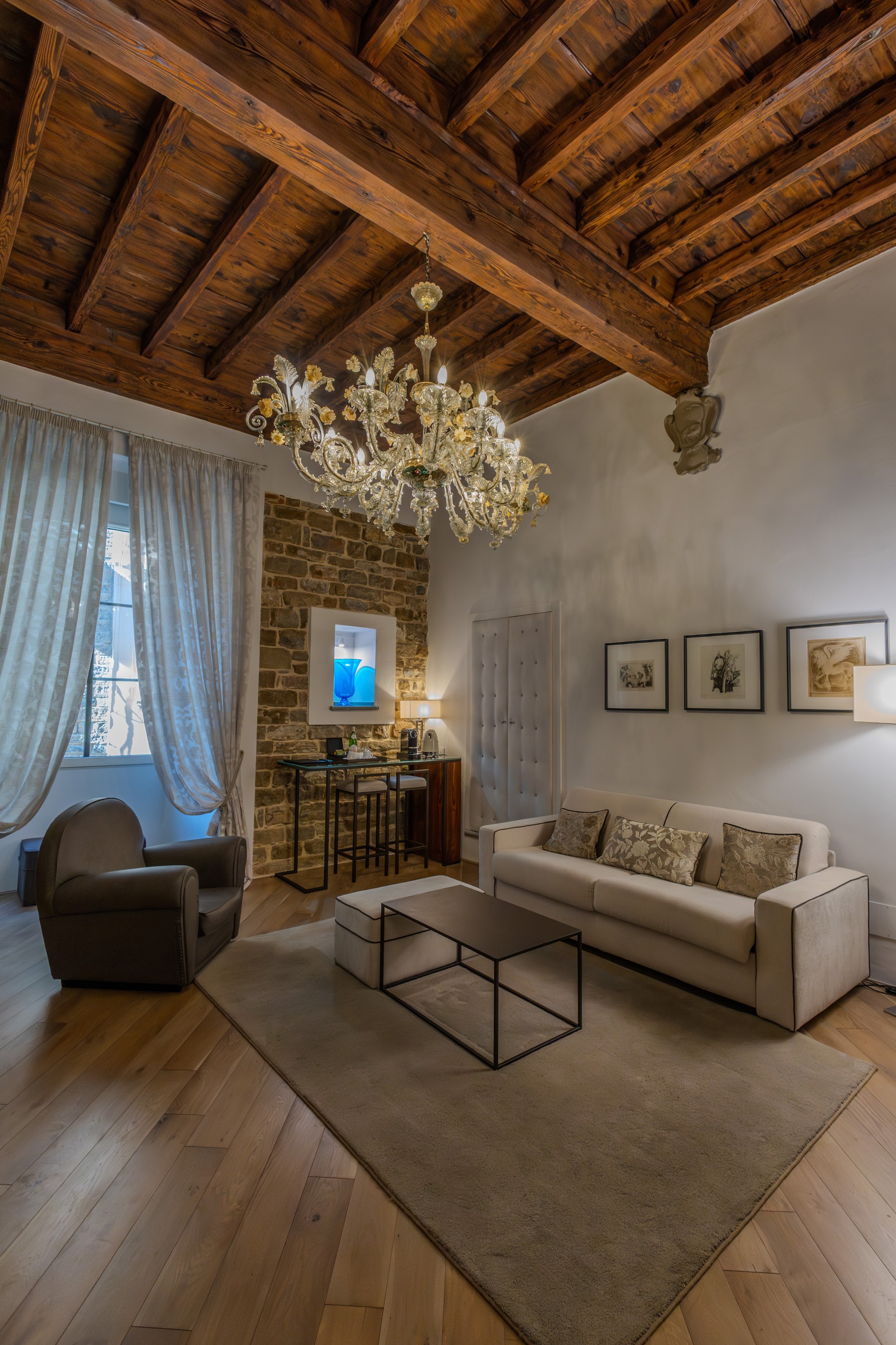 Elegant hotel sitting room featuring exposed wooden beam ceilings, a grand ornate chandelier, cream sofa, armchair, coffee table, and an exposed stone wall with a small bar counter.