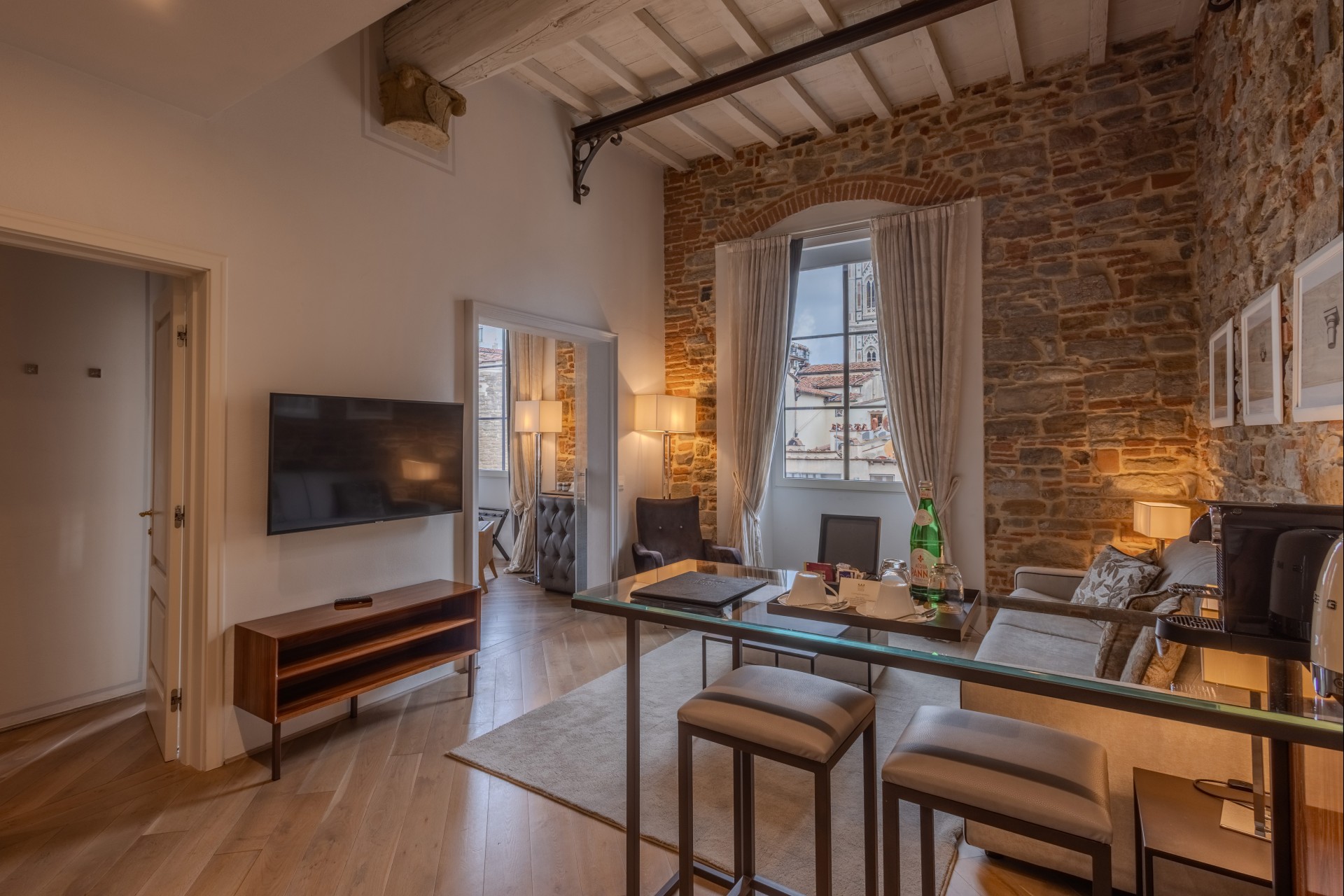 Modern living room area of a luxury suite with exposed stone walls, wooden ceiling beams, high-top bar seating, a sofa, wall-mounted TV, and views of historical architecture through the window.