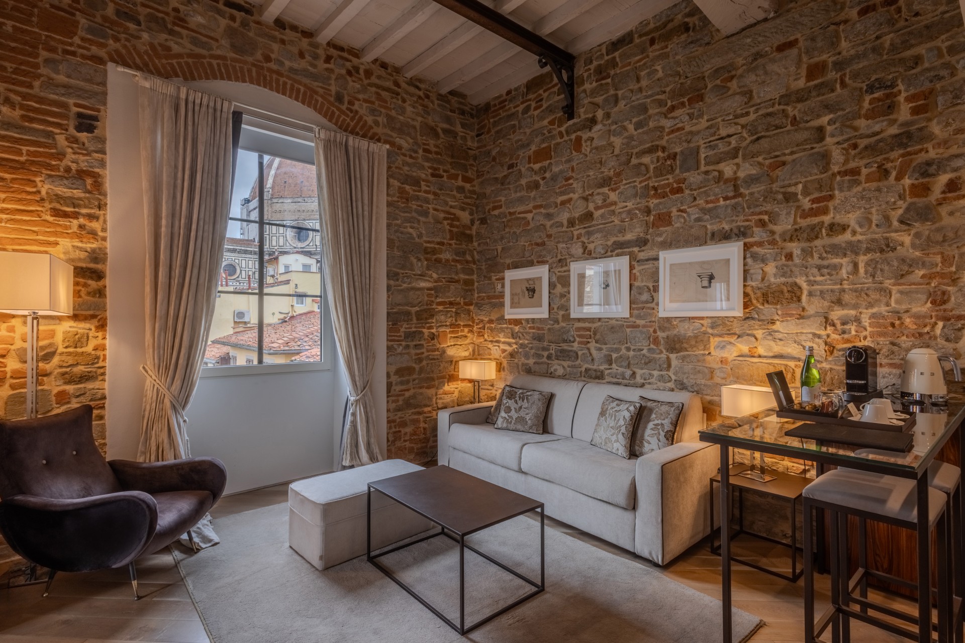 Hotel living area with rustic brick and stone walls, a light grey sofa, a dark velvet armchair, and a window framed by curtains showing the dome of the Florence Cathedral.