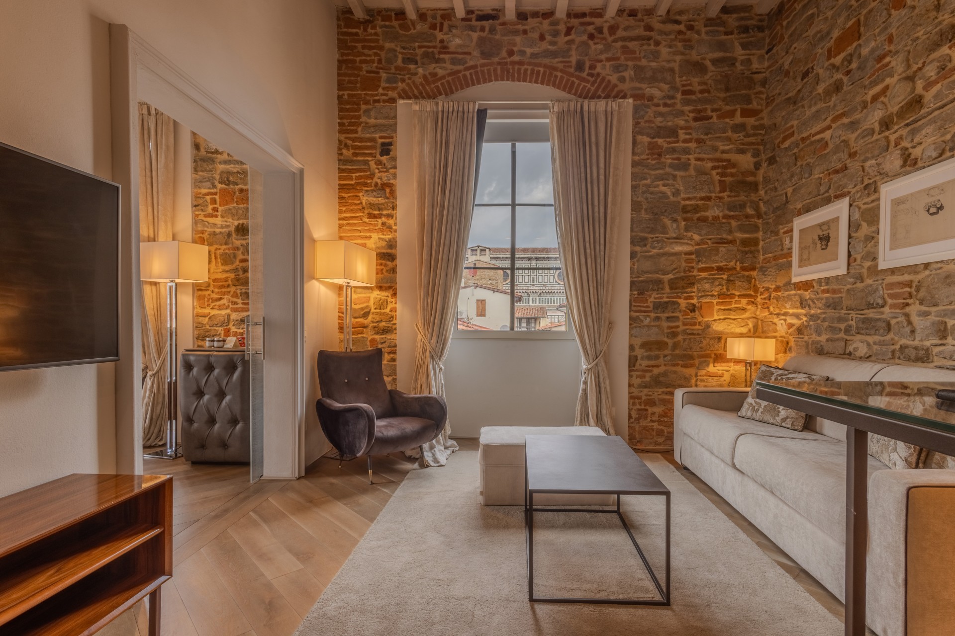 Warmly lit hotel suite sitting area featuring rustic exposed stone walls, a beige sofa, a dark velvet armchair, and a window framed by curtains looking out toward historical Florentine architecture.