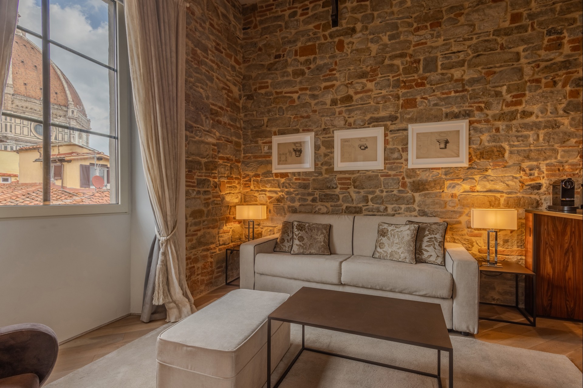Cozy suite living room with an exposed stone wall, cream sofa, dark coffee table, and a window offering a view of the Florence Cathedral dome.