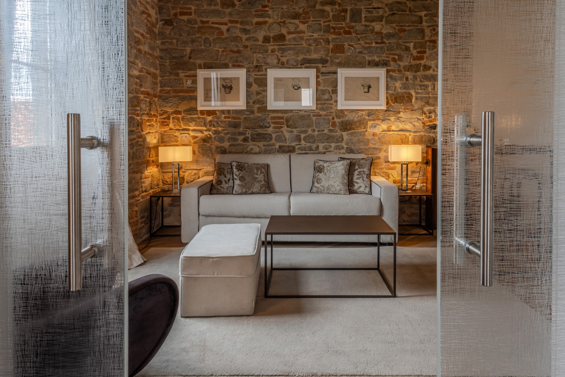 View through glass double doors into a hotel suite living room featuring an exposed stone wall, a beige sofa flanked by lamps, an ottoman, and a minimalist coffee table.