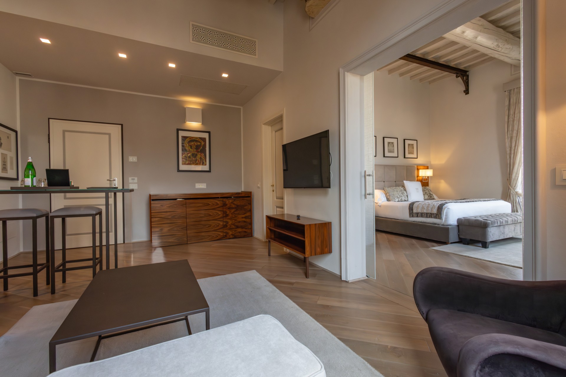 Modern hotel suite living area showing light wood floors, a coffee table, a high table with stools, and an open doorway looking into a warmly lit bedroom.