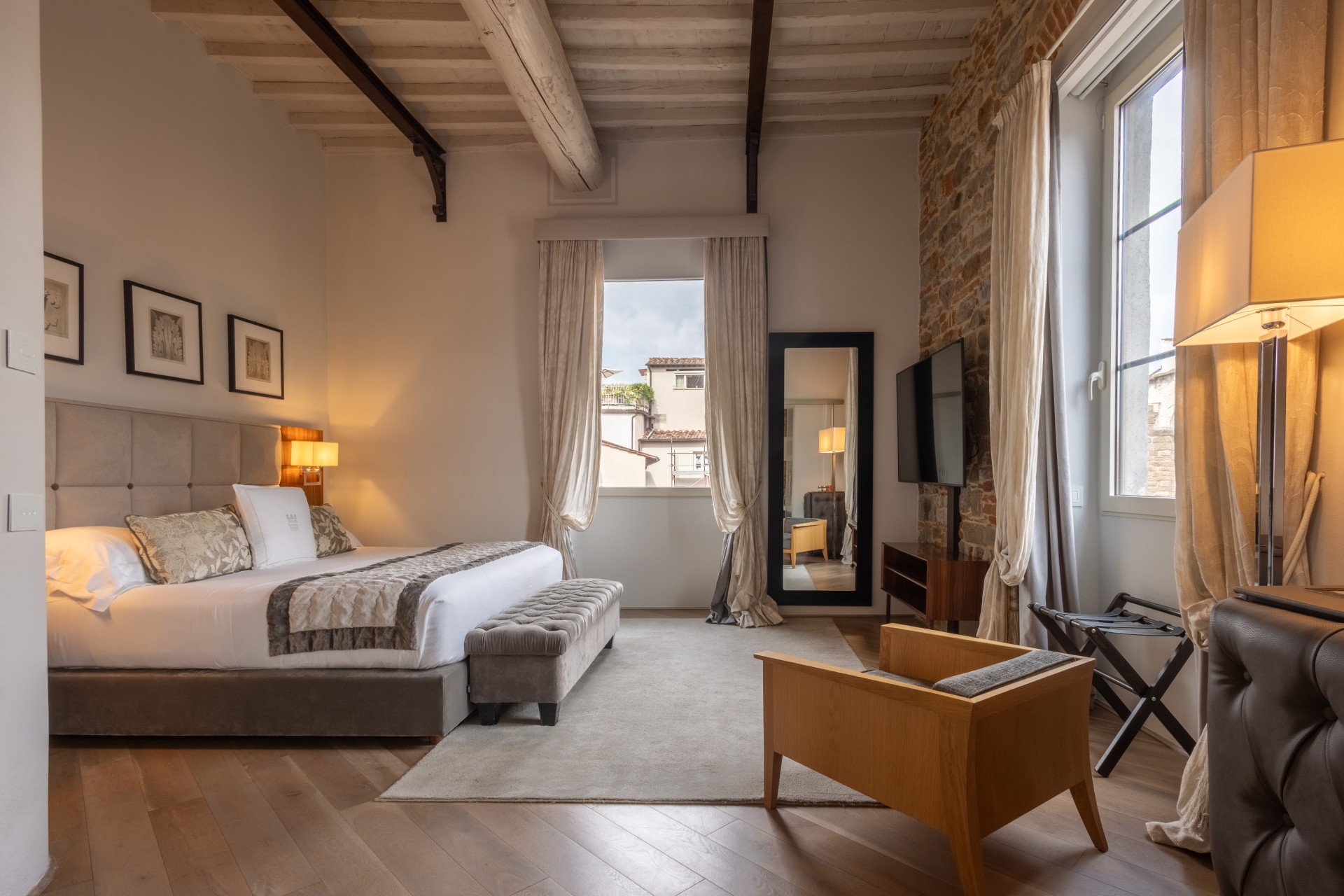 Spacious luxury suite living room with exposed stone walls, a large TV, wooden ceiling beams, an ornate crystal chandelier, and a beige sofa set over a neutral area rug.