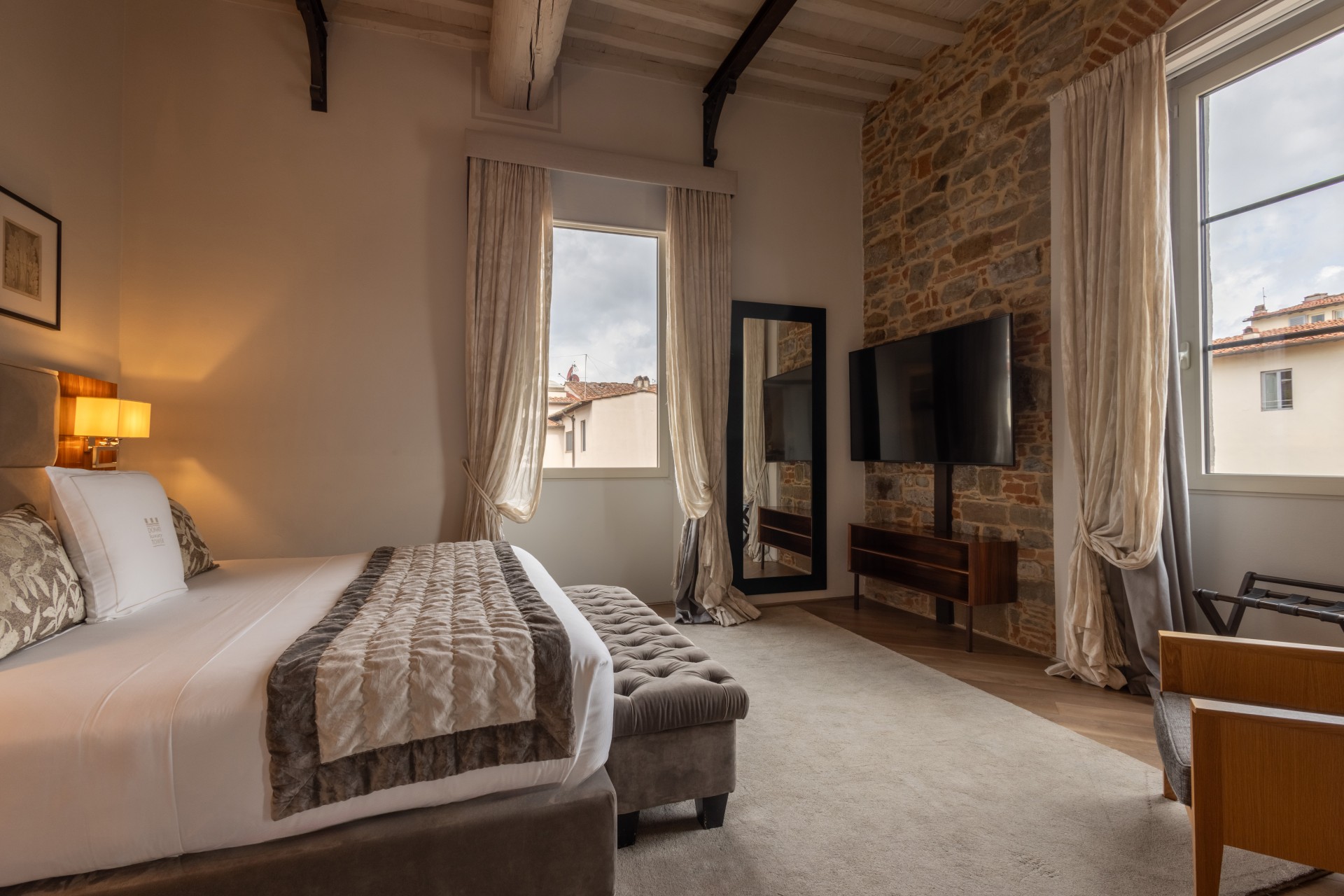 Elegant hotel bedroom featuring a plush bed with a tufted end-of-bed bench, exposed stone wall with a mounted flat-screen TV, high wood-beamed ceilings, and windows looking out onto classic city rooftops.