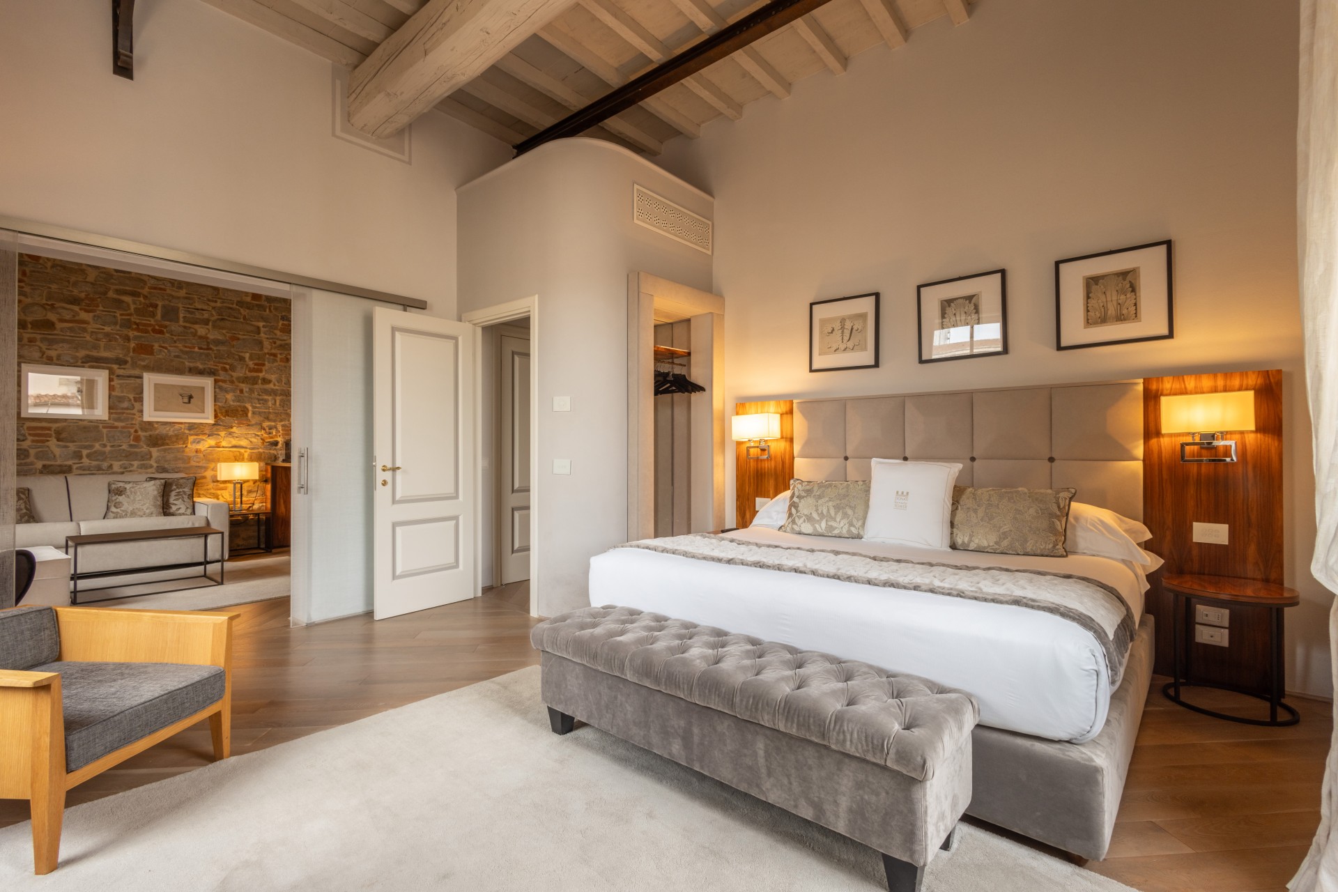 Spacious luxury suite with an upholstered bed, tufted bench, exposed wooden ceiling beams, and an open doorway leading into an adjoining living room area with a stone feature wall.